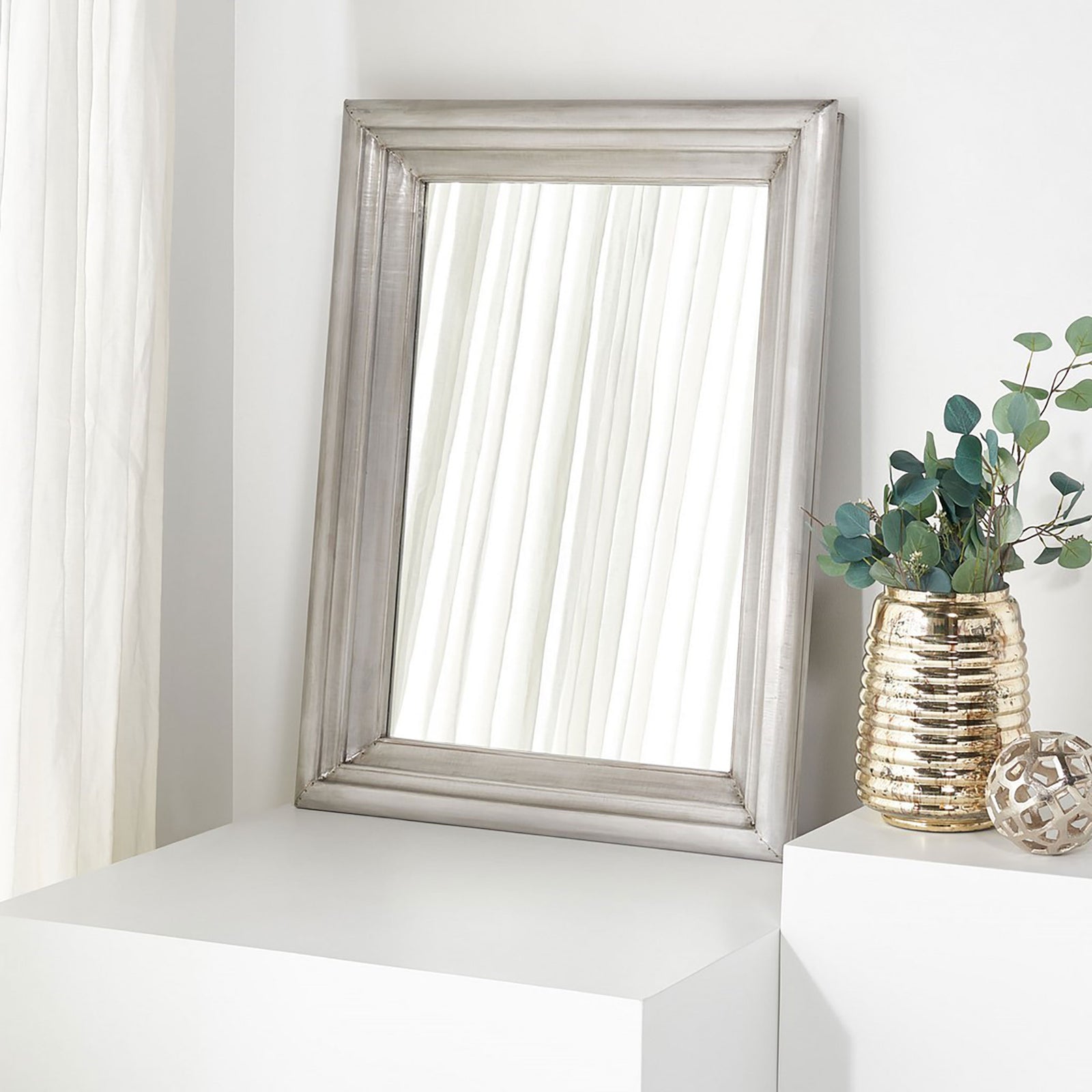 A silver-framed mirror leans against a light wall beside a textured metallic vase. || CMI2002B || front_room