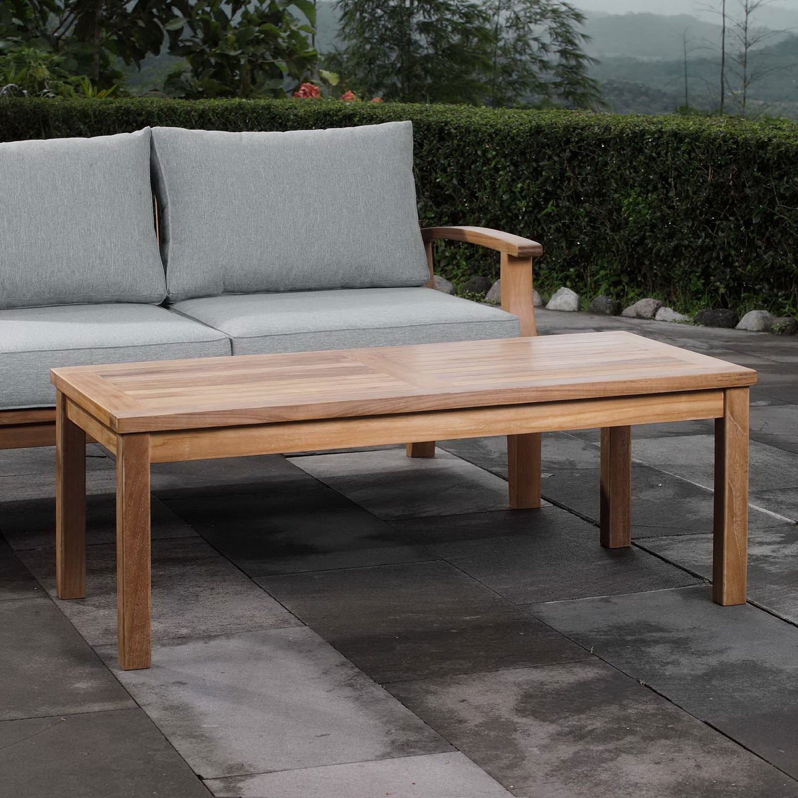 Rectangular teak coffee table styled on a patio between cushions for relaxed outdoor gatherings. || EEI-1154-NAT || front_angle_room