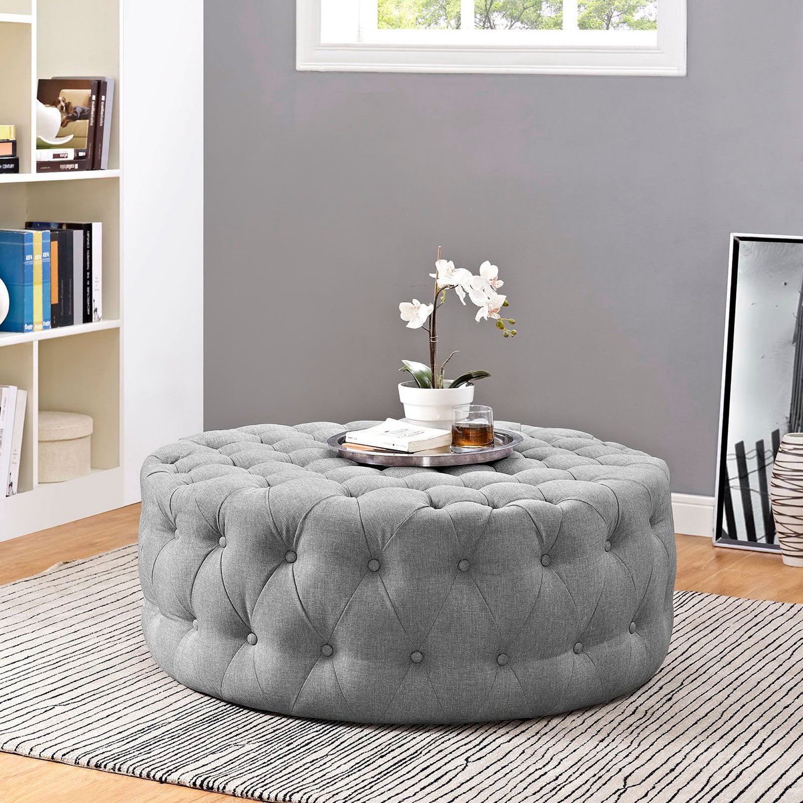 Round tufted ottoman styled as a coffee table with decor on a striped rug || EEI-2225-LGR || front_angle_room
