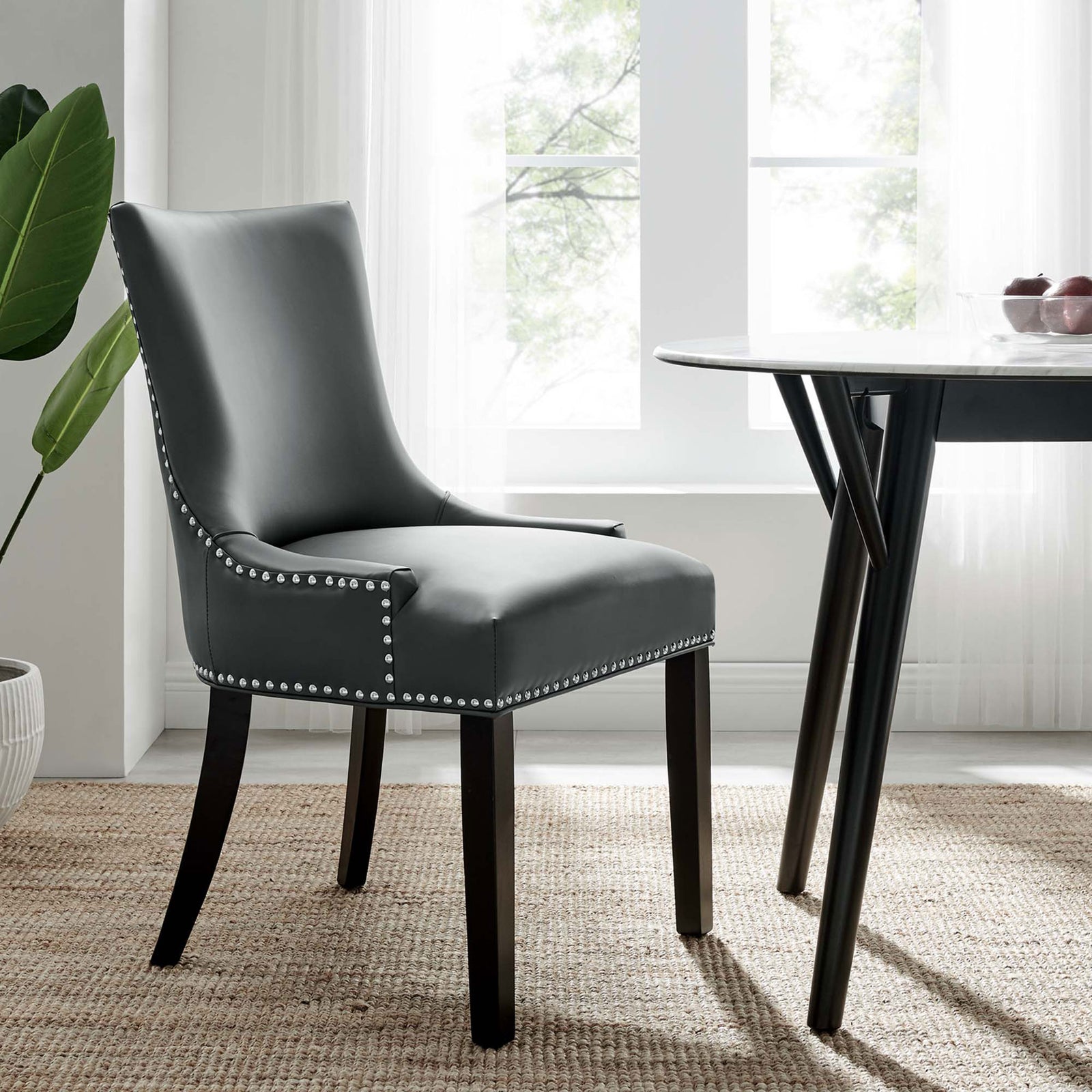 Gray vegan leather dining chair with tall back and nailhead trim beside a modern dining table in a bright room || EEI-2228-GRY || front_angle_room