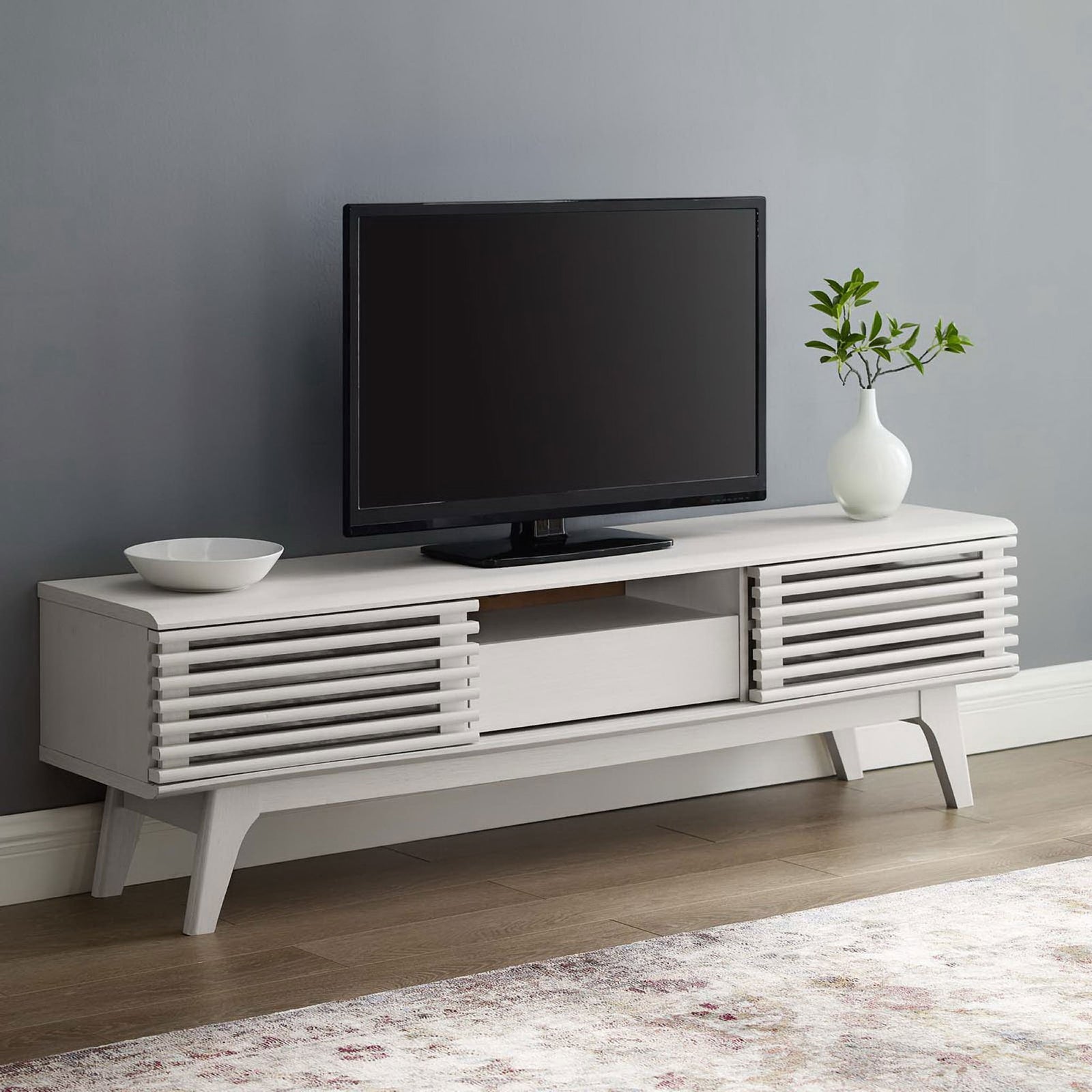White TV stand styled in a modern living room with TV, rug, and simple decor || EEI-2541-WHI || front_angle_room