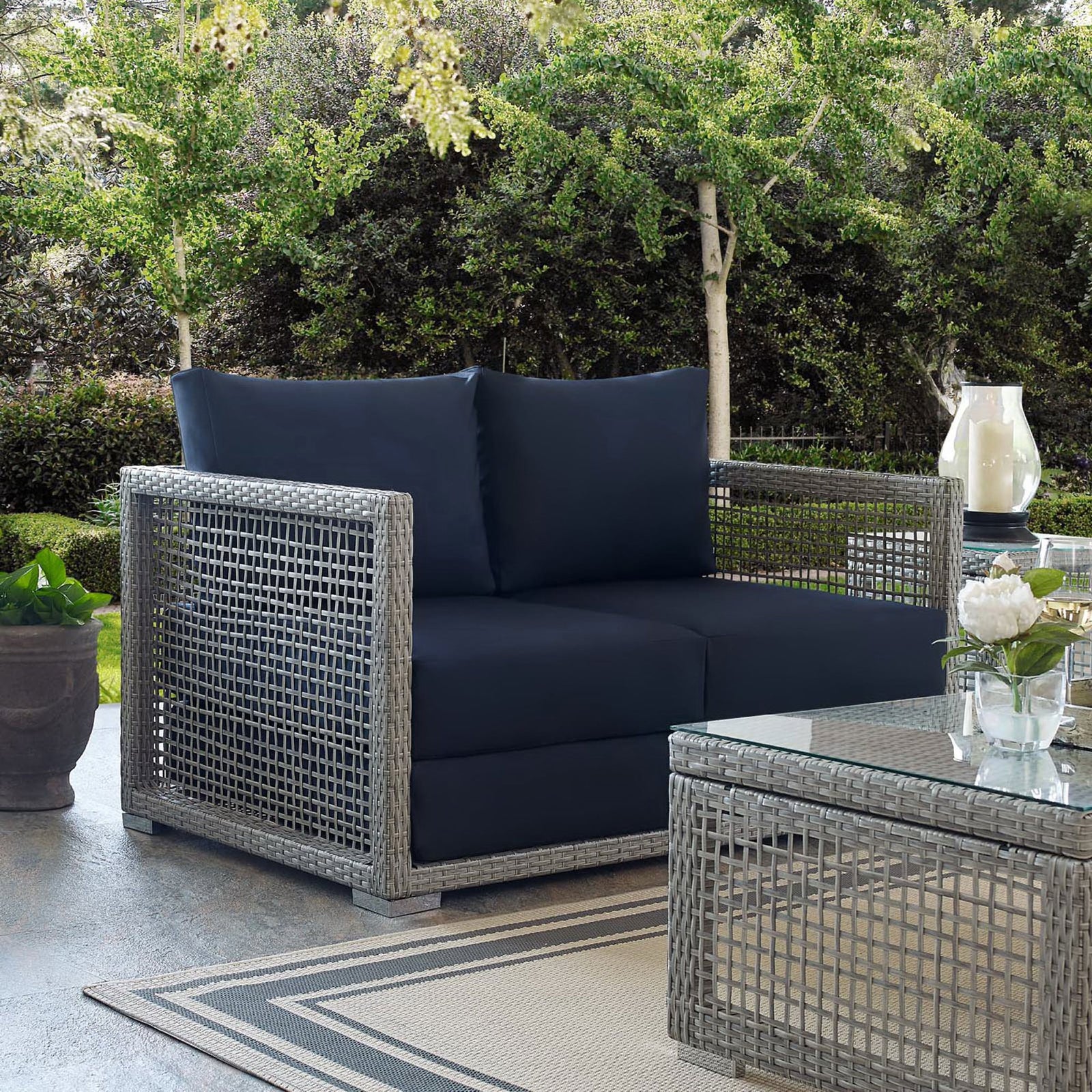 Gray wicker loveseat with navy cushions styled on a lush outdoor patio || EEI-2924-GRY-NAV || front_angle_room