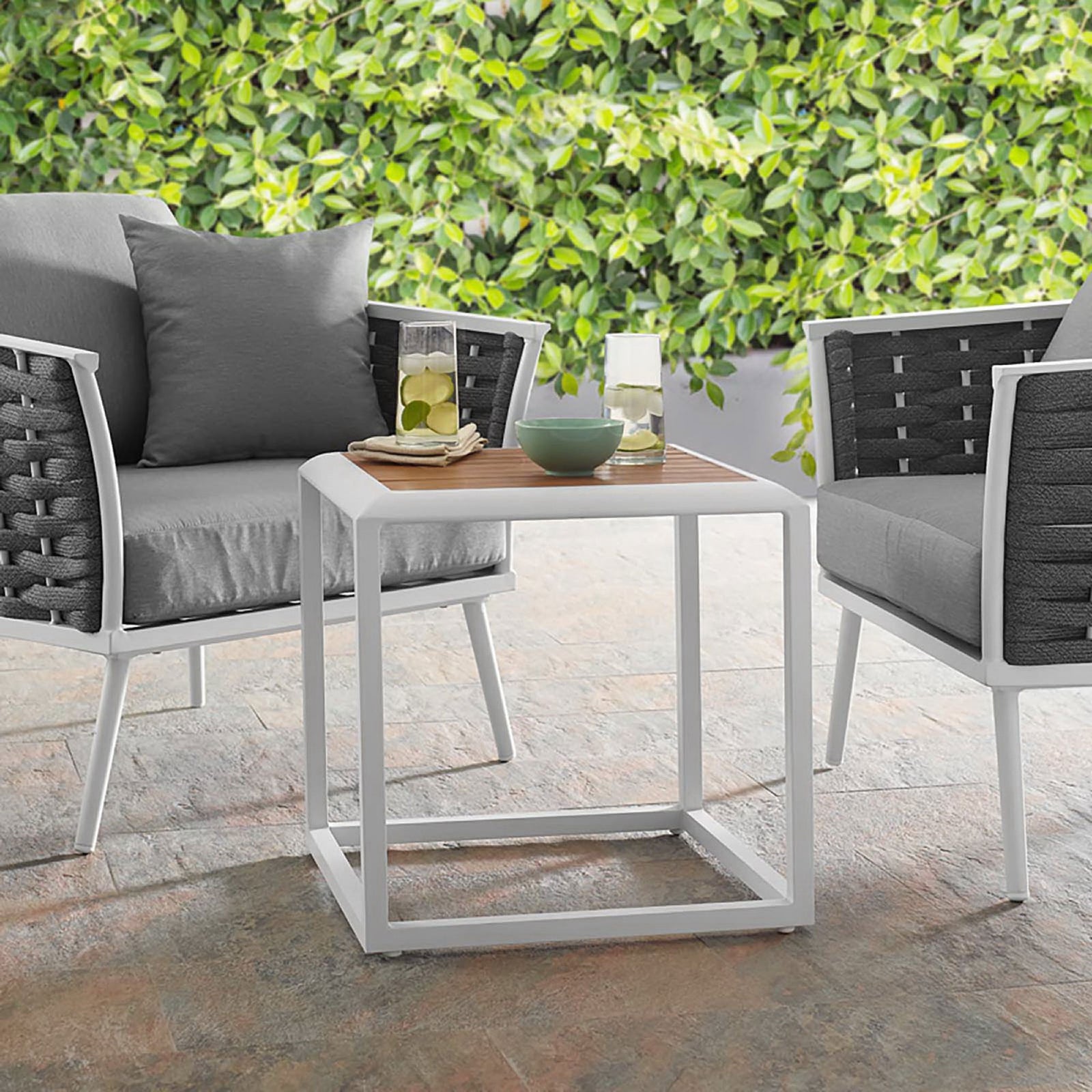 Outdoor side table styled between cushioned lounge chairs on a stone patio with greenery || EEI-3022-WHI-NAT || front_angle_room