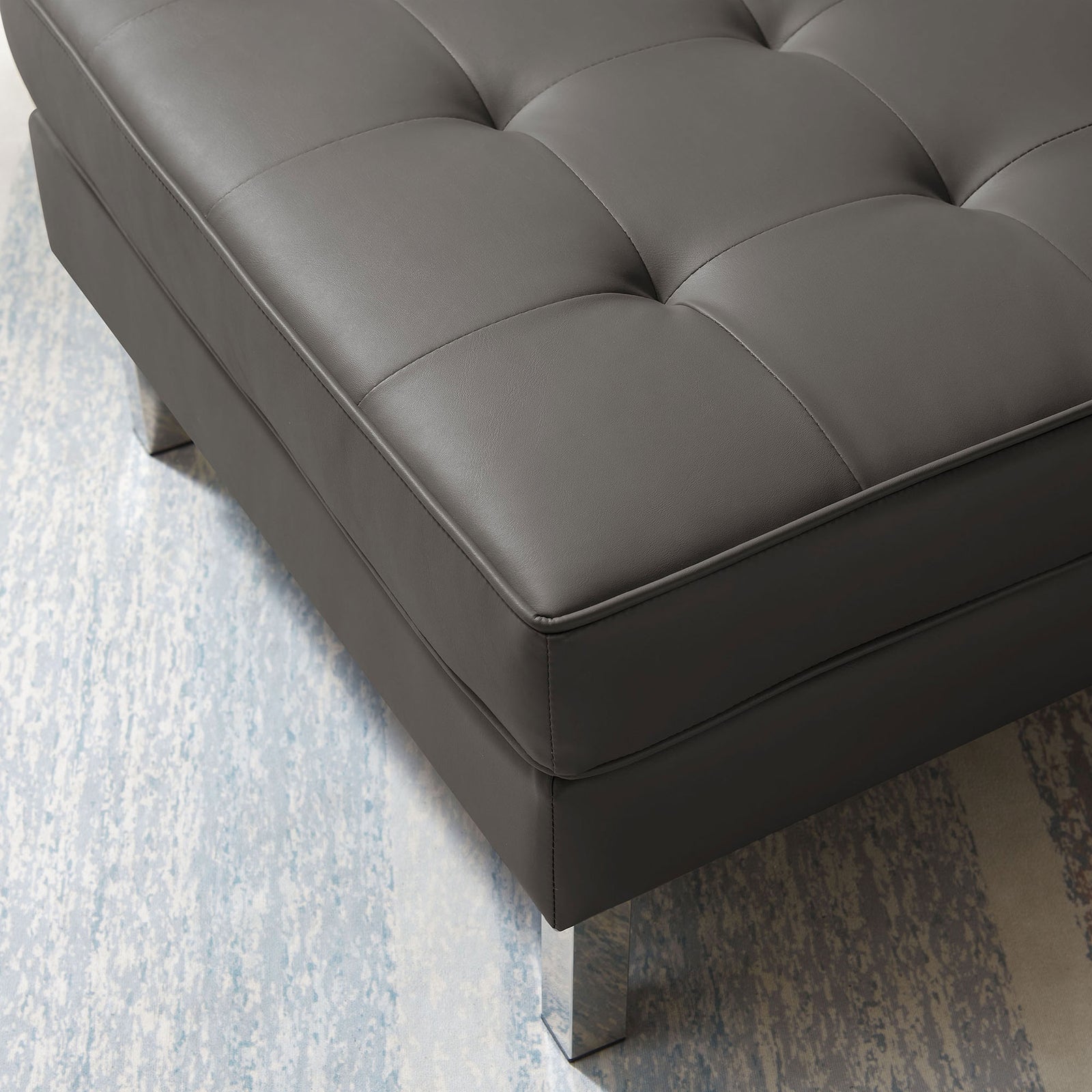 Detail of silver gray vegan leather ottoman corner showing biscuit tufting and shiny leg over a patterned rug || EEI-3394-SLV-GRY || detail_room