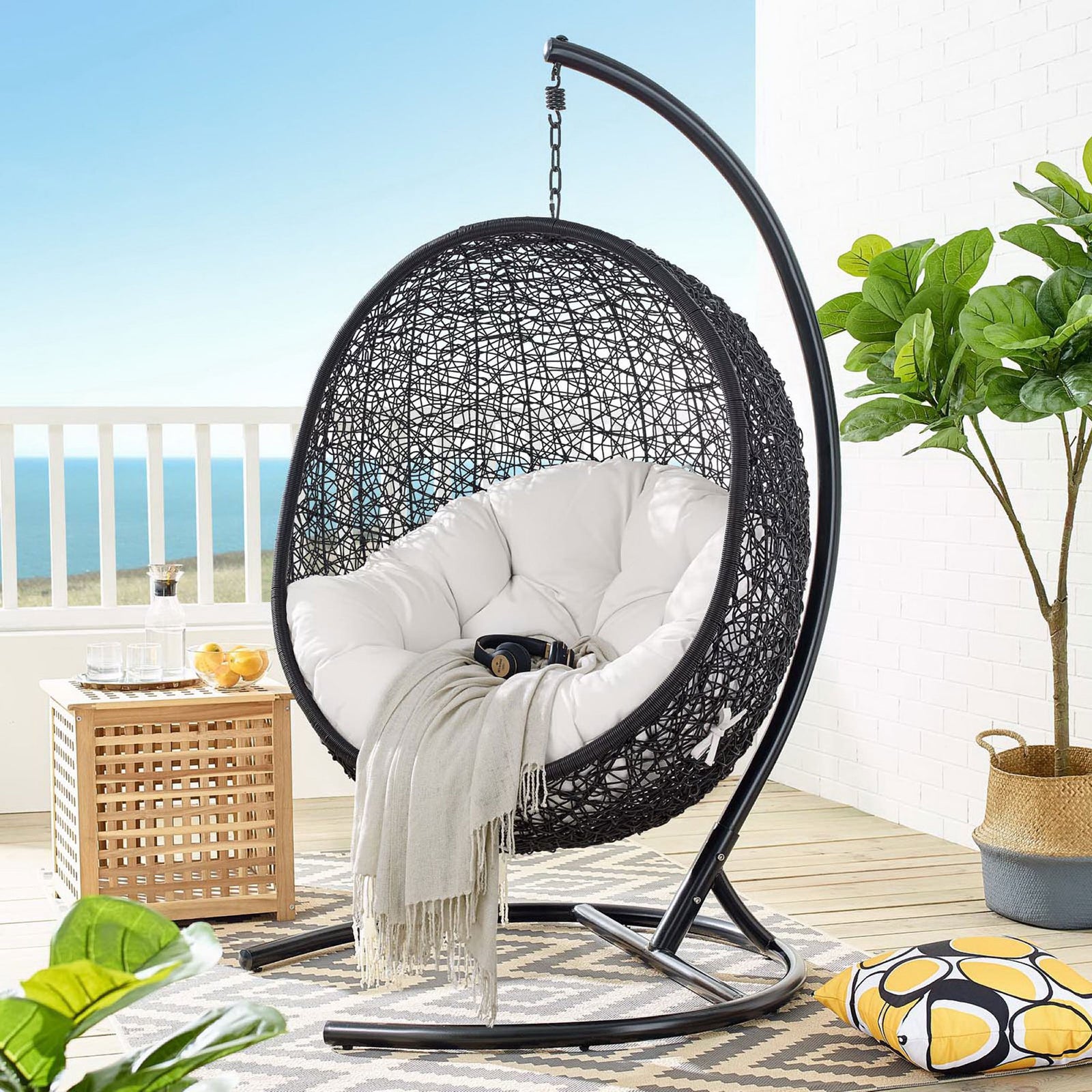 White cushioned hanging egg chair and stand styled on a bright balcony with rug and greenery || EEI-3943-BLK-WHI || front_angle_room