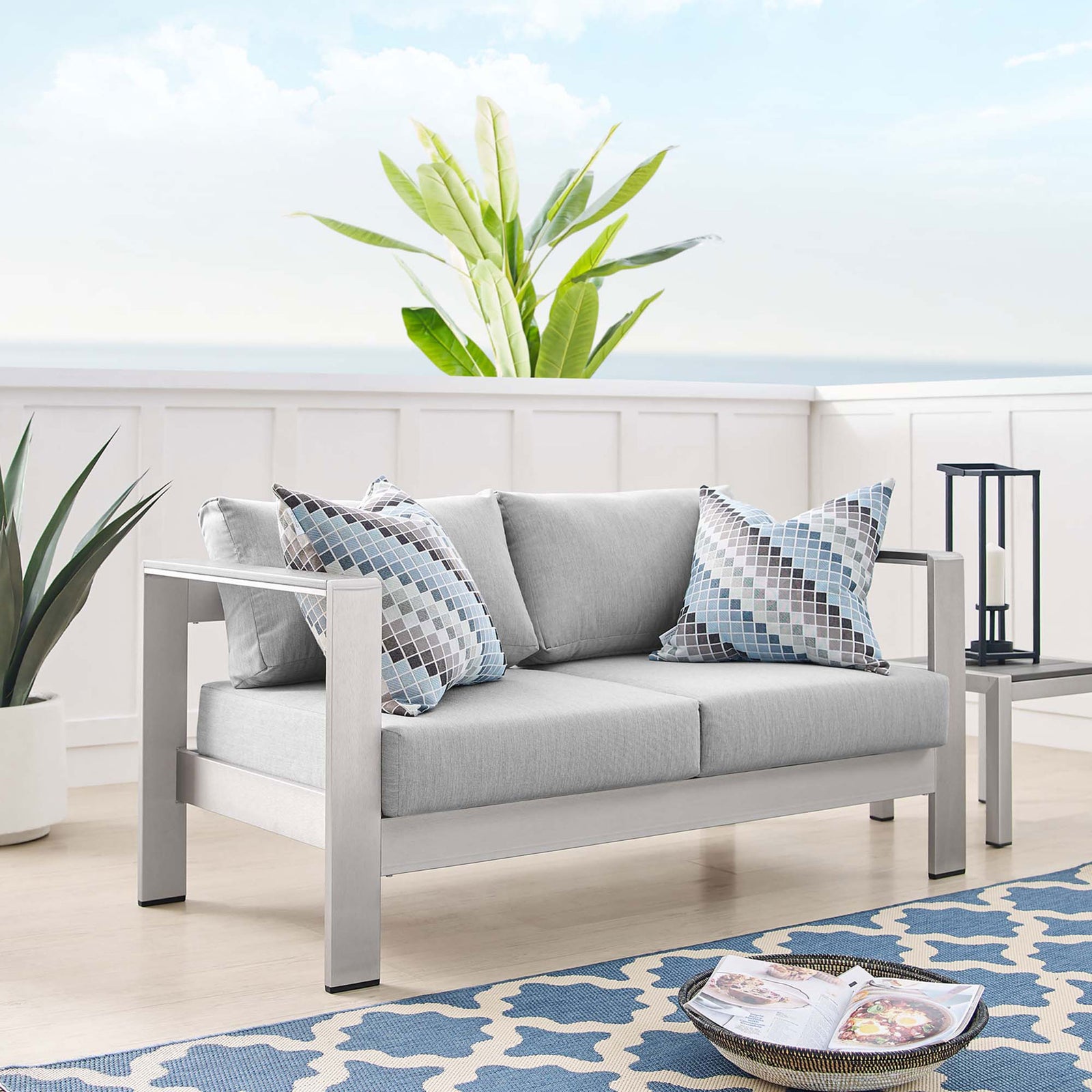 Light gray cushioned aluminum loveseat styled with patterned pillows in a bright coastal-inspired room || EEI-4226-SLV-GRY || front_angle_room
