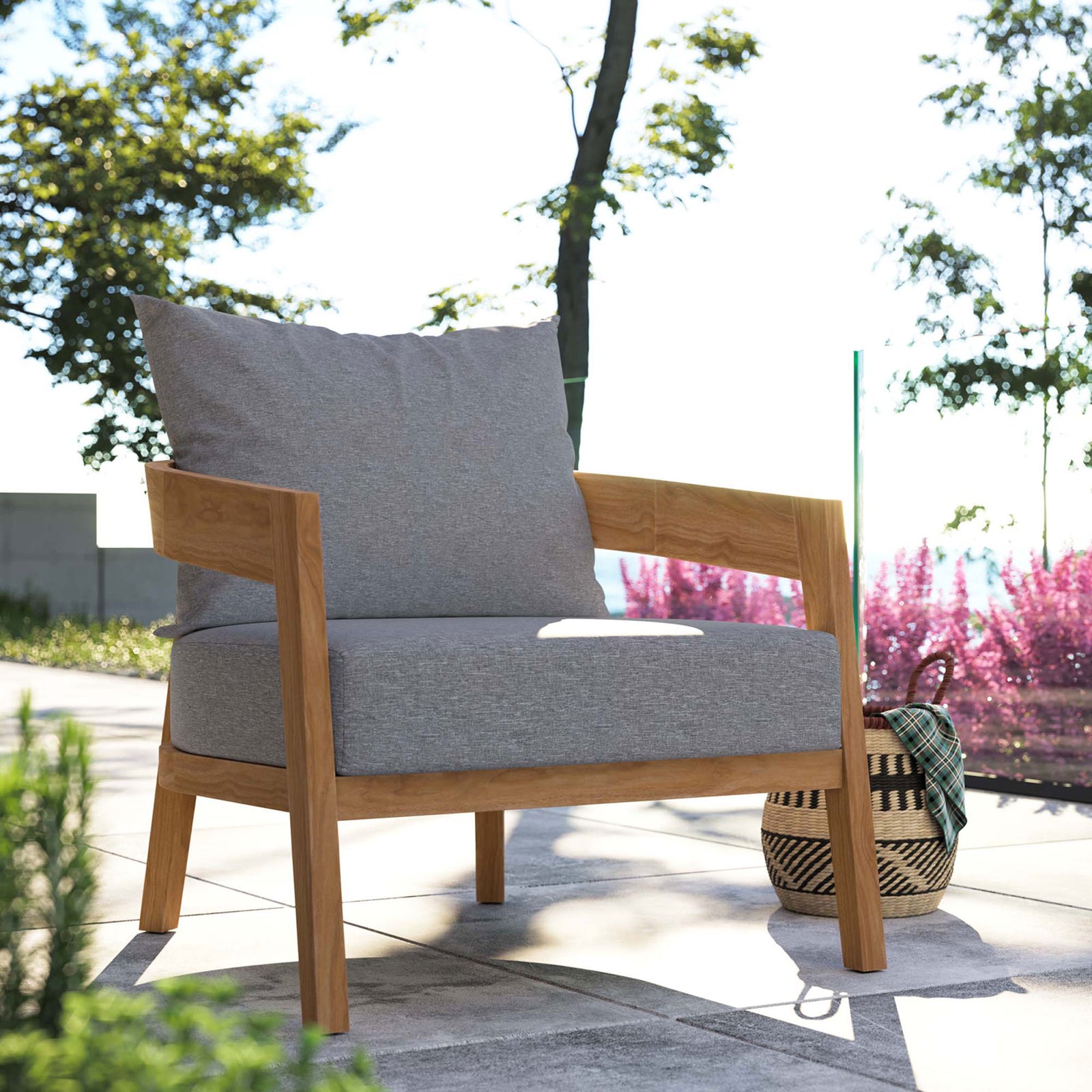 Gray cushioned teak outdoor armchair on a sunlit patio surrounded by greenery and bright flowering plants || EEI-5602-NAT-GRY || front_angle_room
