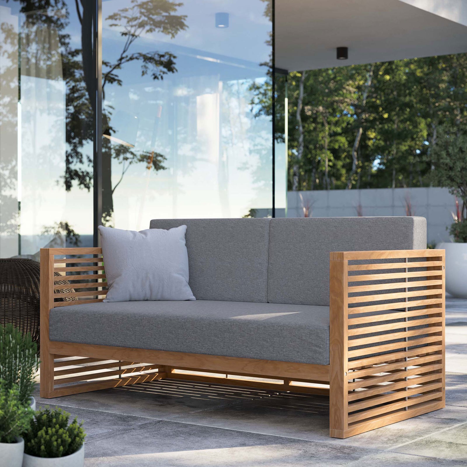 Carlsbad teak outdoor loveseat with gray cushions styled on a modern patio with lush greenery views. || EEI-5605-NAT-GRY || front_angle_room