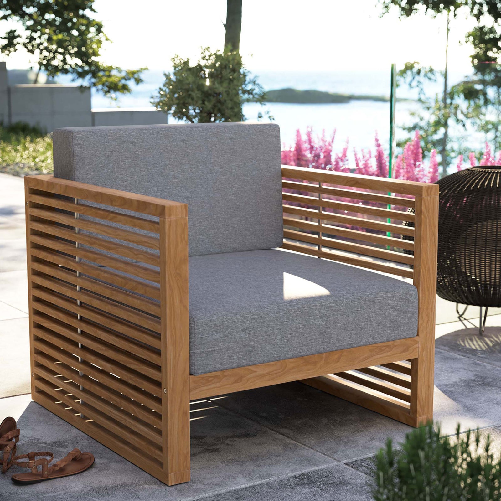 Carlsbad teak outdoor armchair with gray cushions styled on a sunny stone patio overlooking water. || EEI-5606-NAT-GRY || front_angle_room