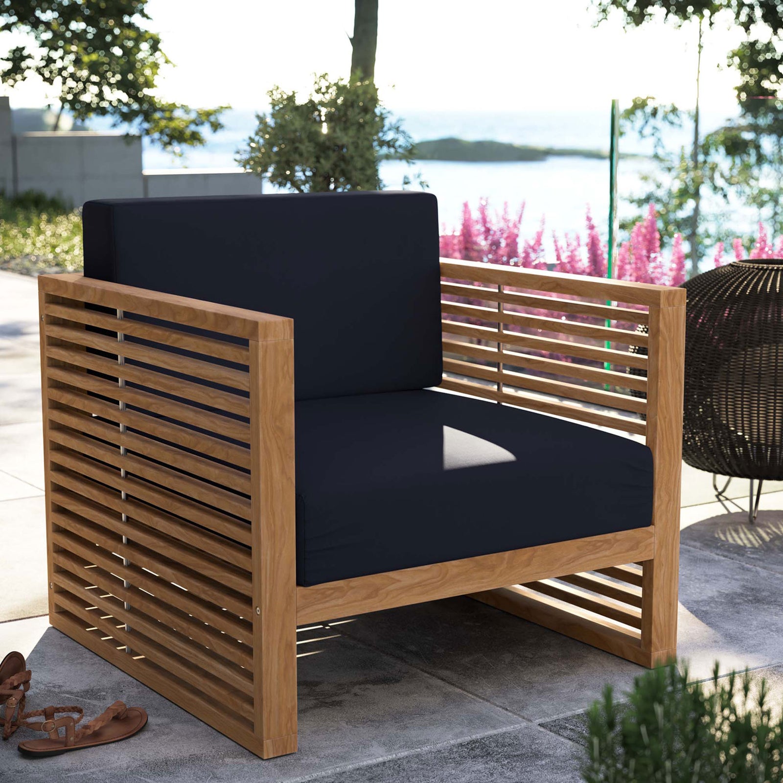 Carlsbad teak outdoor armchair with navy cushions arranged on a terrace overlooking the water. || EEI-5606-NAT-NAV || front_angle_room