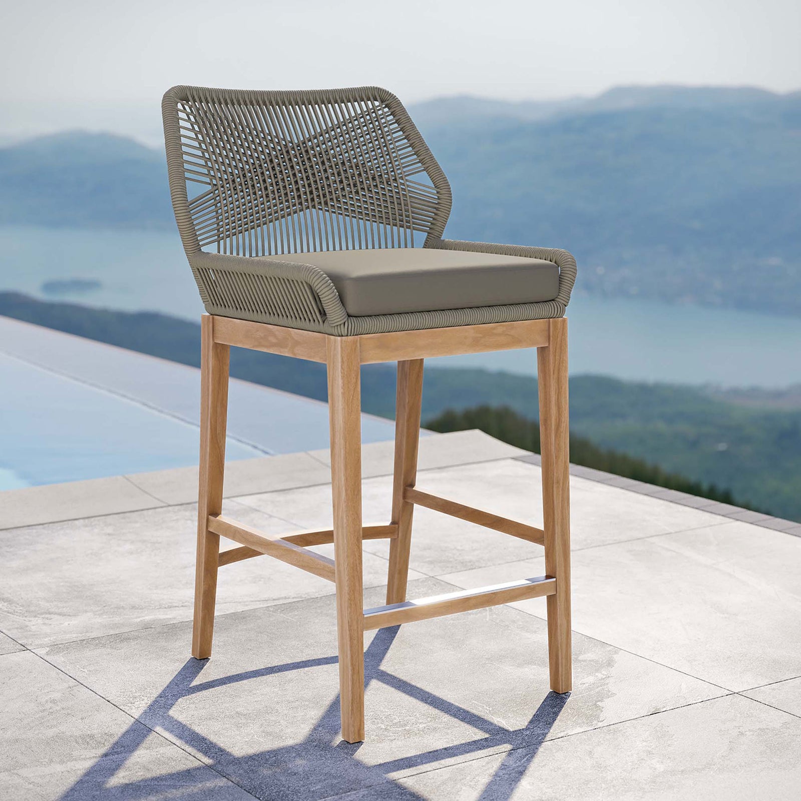 Light gray greige rope bar stool with cushioned seat and teak base styled on a serene poolside patio. || EEI-5746-LGR-GRG || front_angle_room