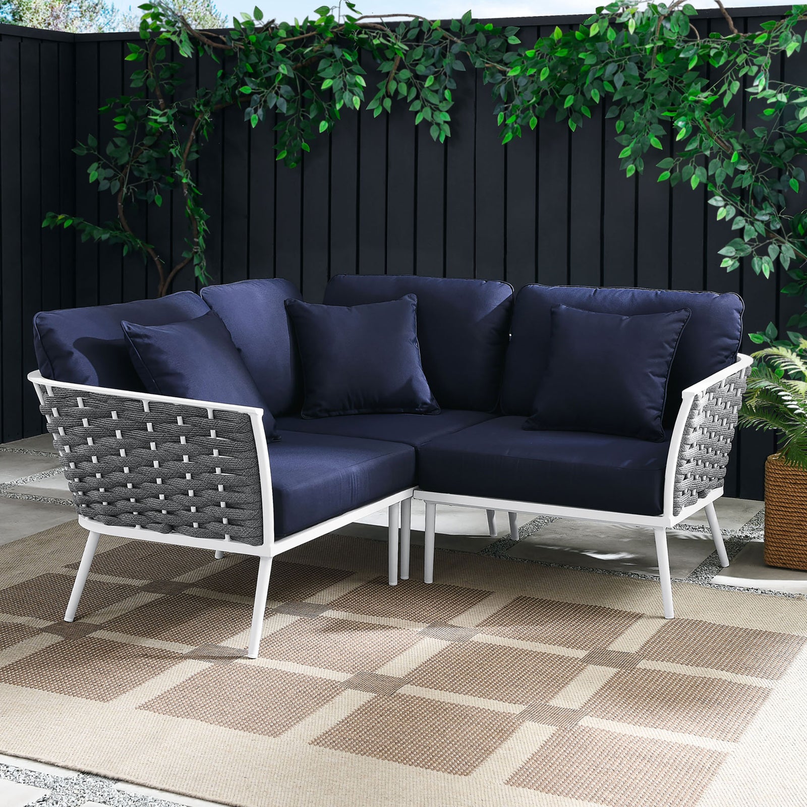 Navy cushion sectional anchors a modern patio with white frame, taupe outdoor rug, and lush greenery || EEI-5752-WHI-NAV || front_angle_room