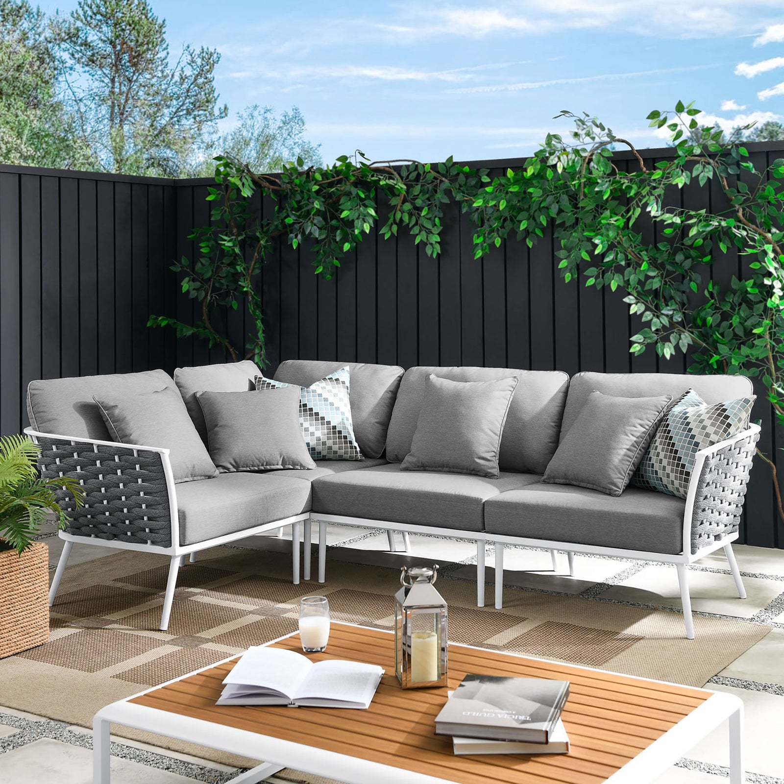Gray outdoor sectional anchors a modern patio lounge with lush greenery, accent pillows, and a coordinating coffee table. || EEI-5753-WHI-GRY || front_angle_room