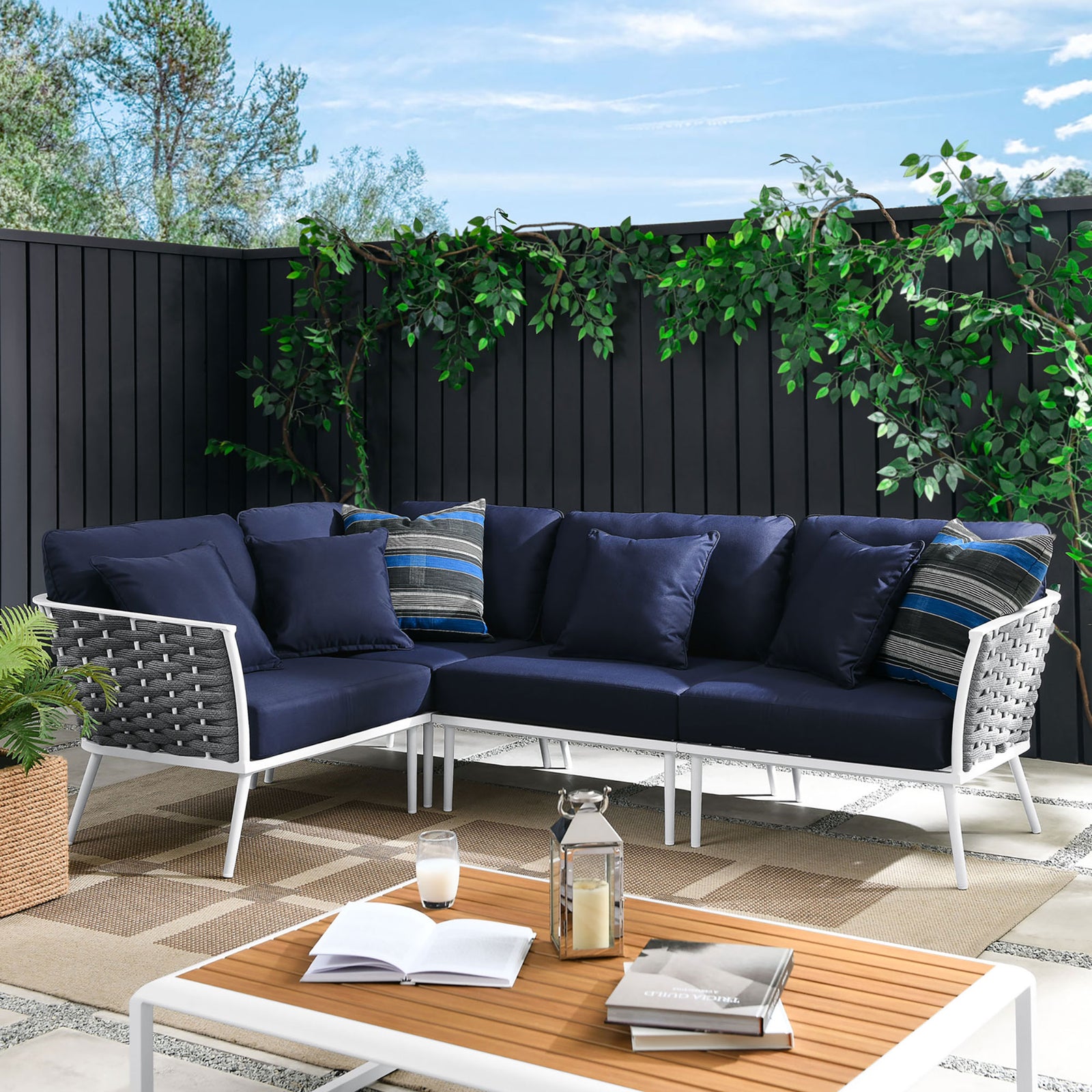 Navy outdoor sectional creates an inviting patio lounge framed by greenery, a wood coffee table, and layered rugs. || EEI-5753-WHI-NAV || front_angle_room