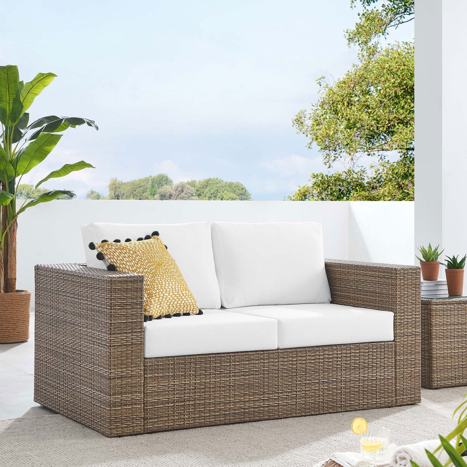 Cappuccino outdoor loveseat with white cushions anchors a bright patio scene with lush greenery and potted plants || EEI-6246-CAP-WHI || front_angle_room