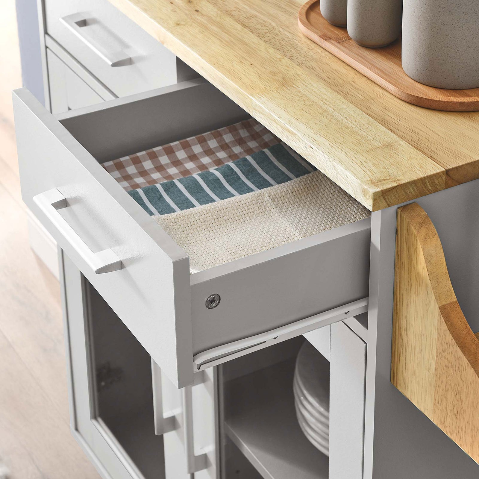 Open drawer of kitchen cart neatly holds folded linens beside styled serveware on the wood top || EEI-6276-LGR-NAT || storage_open_room