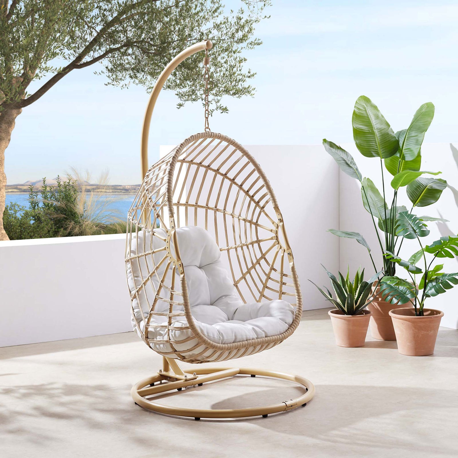 Natural rattan hanging egg chair with stand and white cushion set on a modern patio with tropical plants and ocean view || EEI-6337-NAT-WHI || front_angle_room