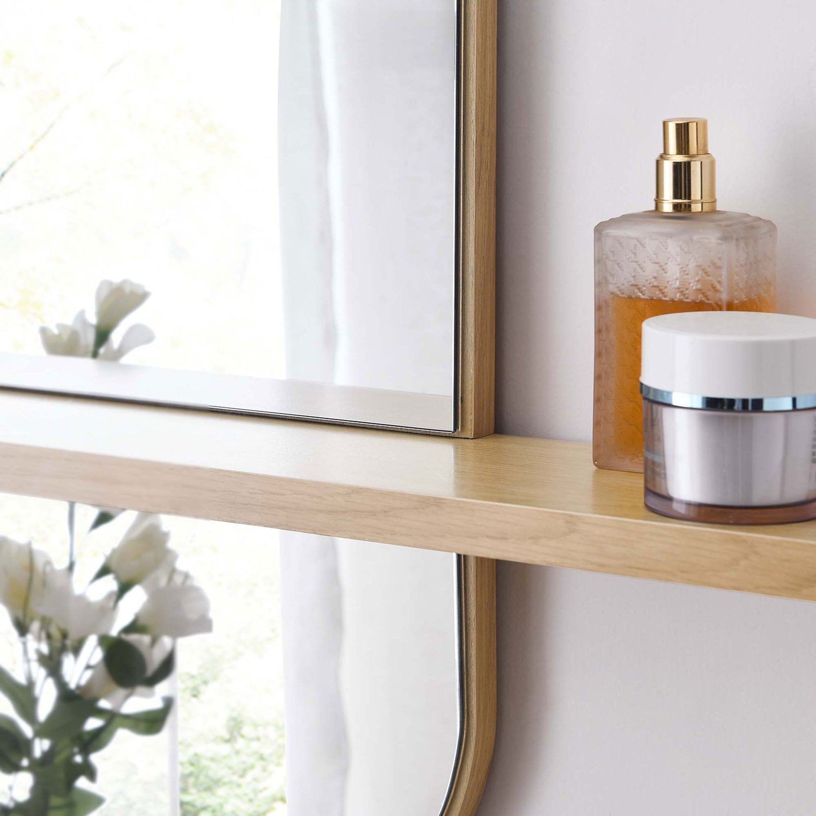 Oak mirror shelf holding skincare bottles beside soft florals and window light. || EEI-6349-OAK || storage_room