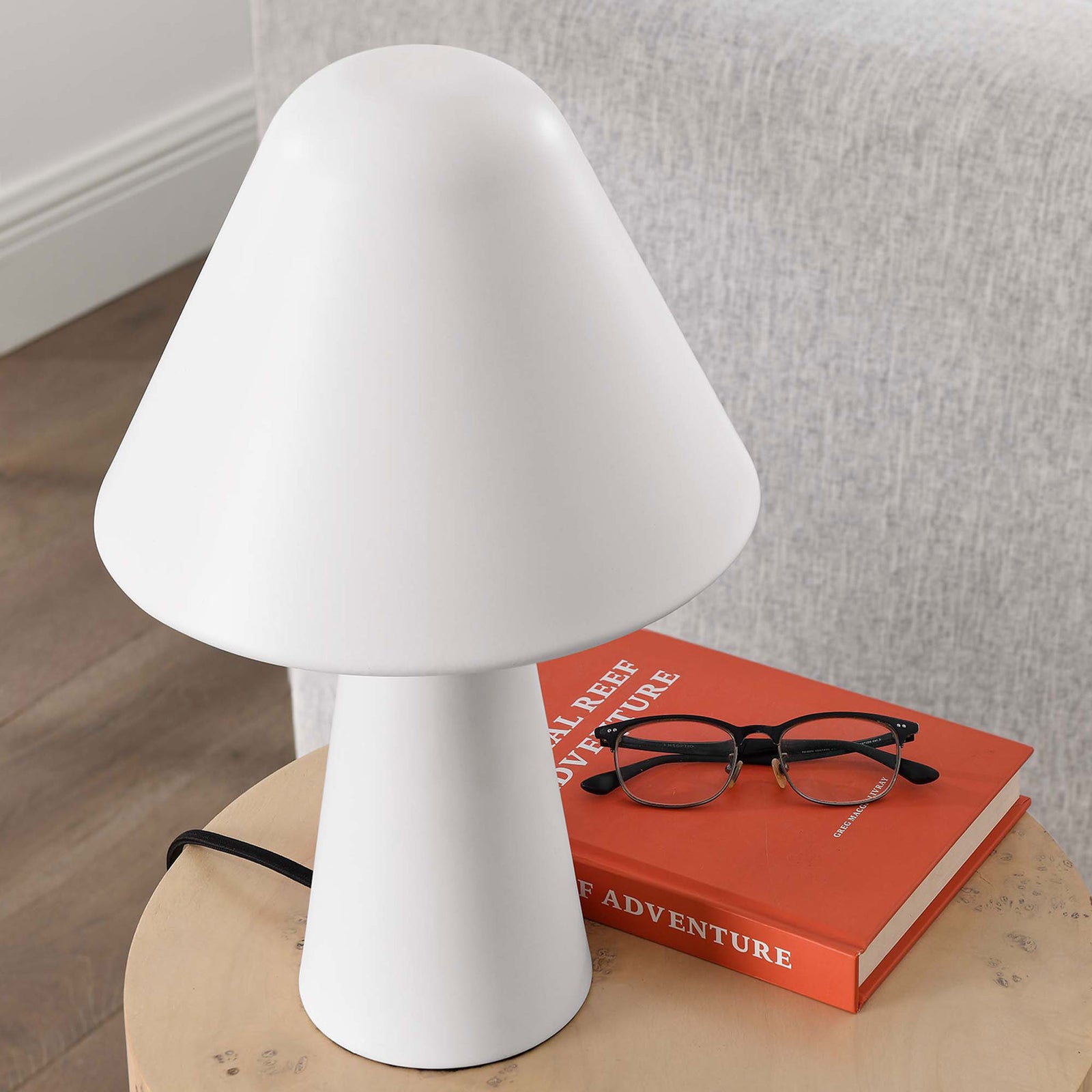 White Jovial mushroom lamp rests on a wood side table beside books and glasses in a lounge. || EEI-6529-WHI || top_angle_room