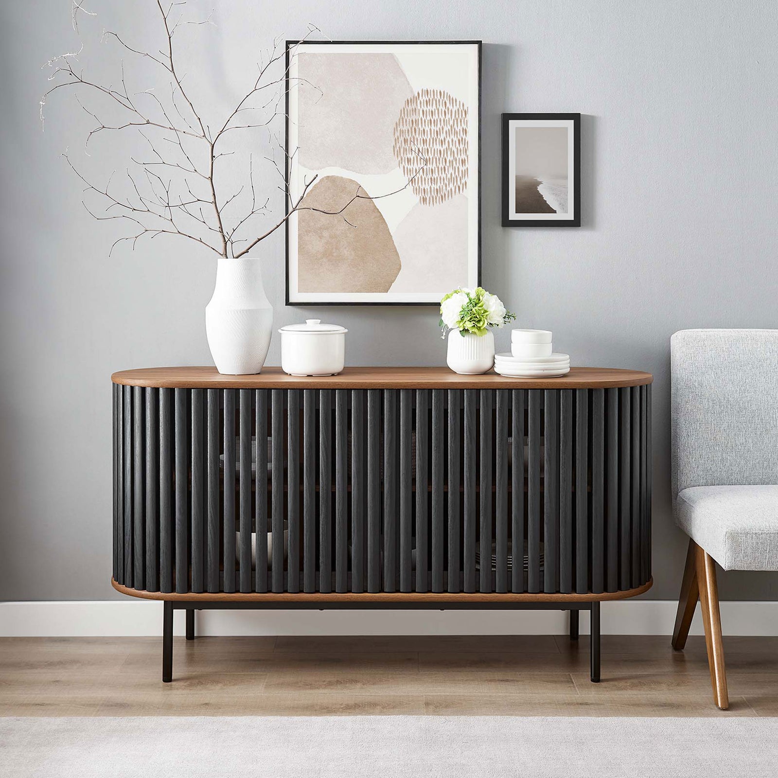 Walnut and black oval sideboard styled in a contemporary room with vases and abstract wall art. || EEI-6590-WAL-BLK || front_room