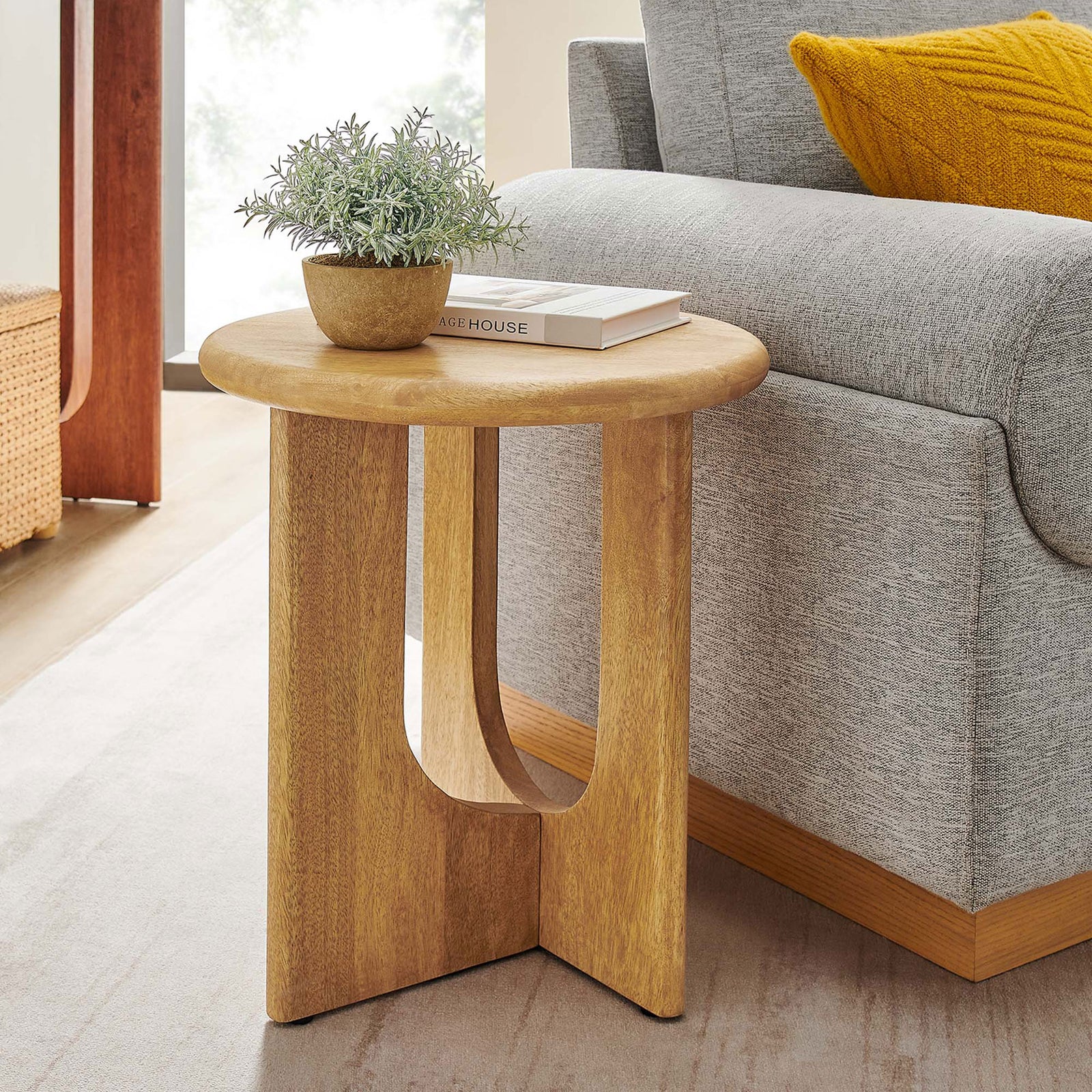 Round oak side table styled beside a modern sofa with decor accents in a bright living room. || EEI-6595-OAK || front_angle_room