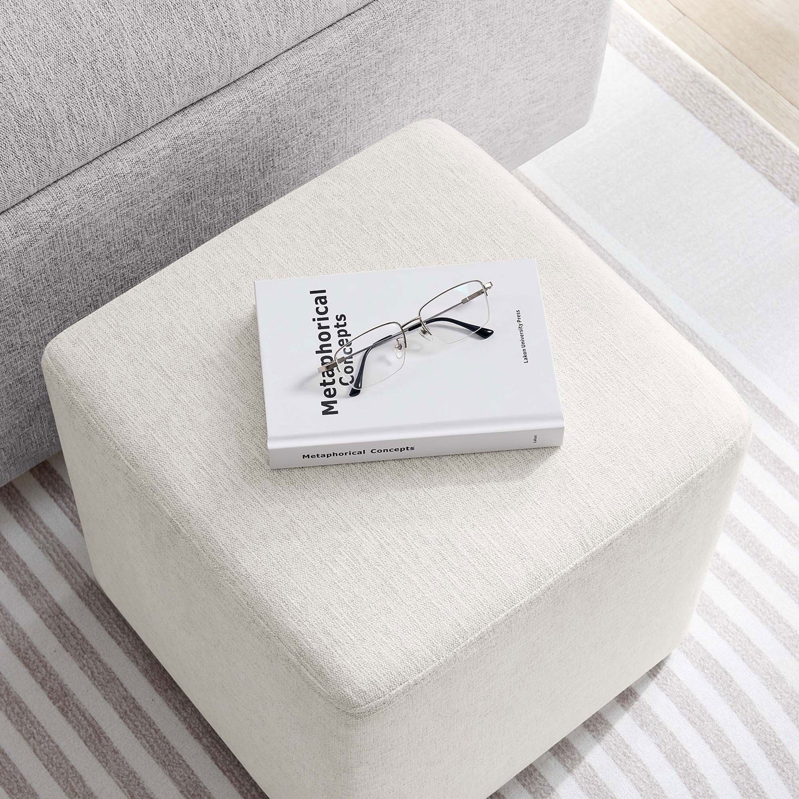Ivory upholstered ottoman styled as a small side table holding a book and glasses in a bright room. || EEI-6636-HEI || top_angle_room