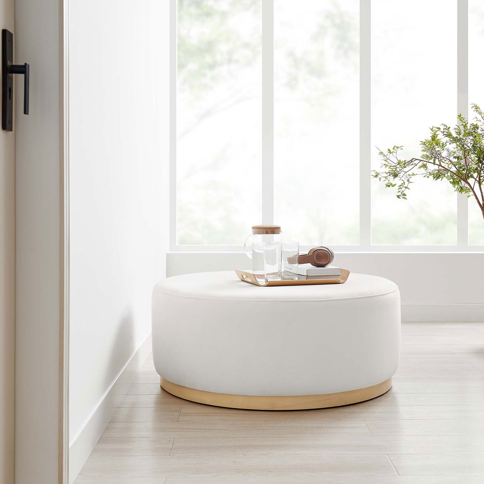 Tilden 38 inch round velvet ottoman used as a casual table in a bright minimalist room with greenery || EEI-6650-ALA-NAT || front_angle_room