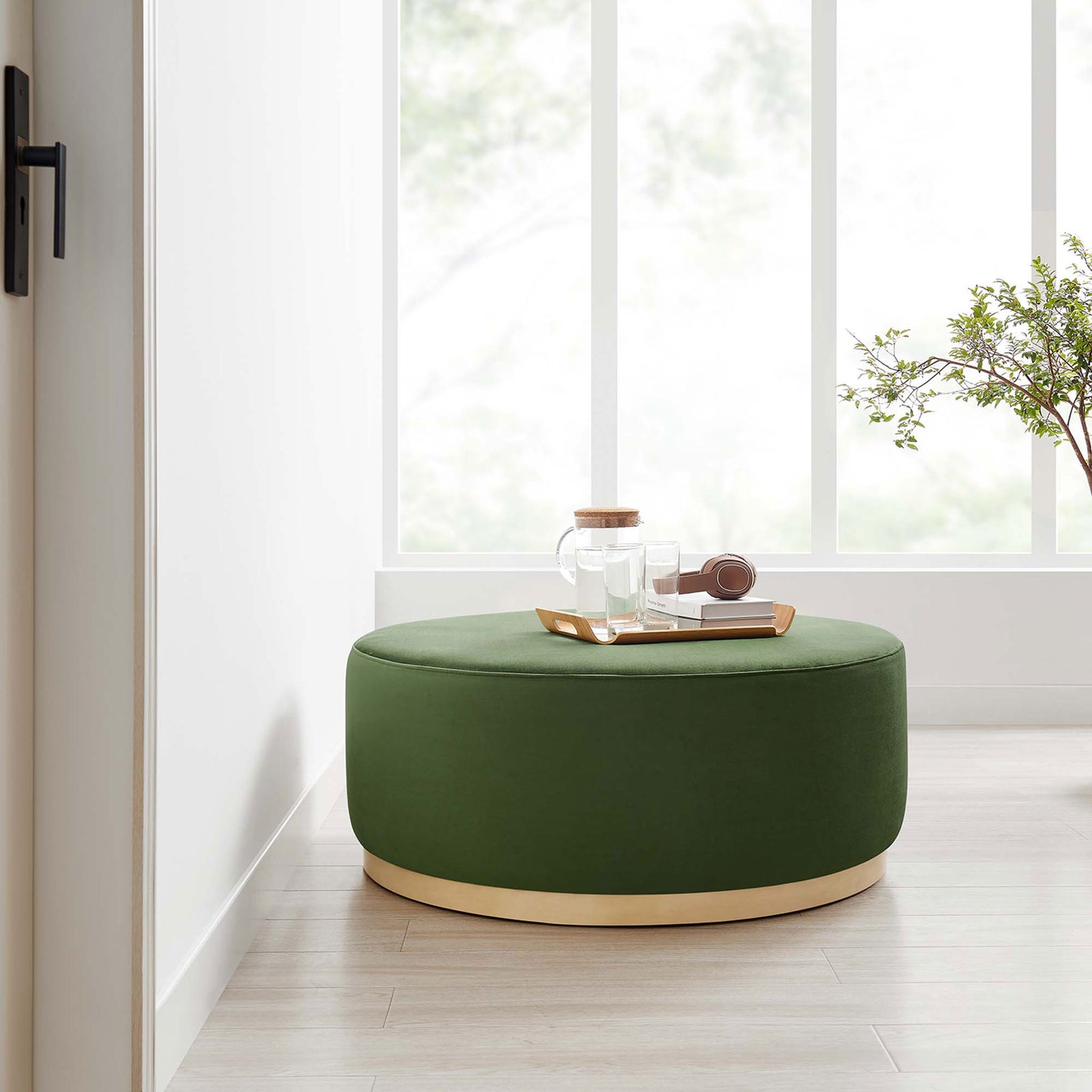Generous moss green velvet round ottoman with wood base serving as a coffee table in a bright airy room. || EEI-6650-MOG-NAT || front_angle_room