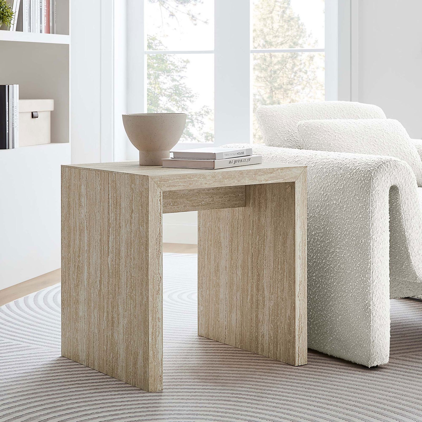Mirella faux travertine side table paired with a curved sofa and round rug for a bright, modern living room || EEI-6772-TRA || front_angle_room
