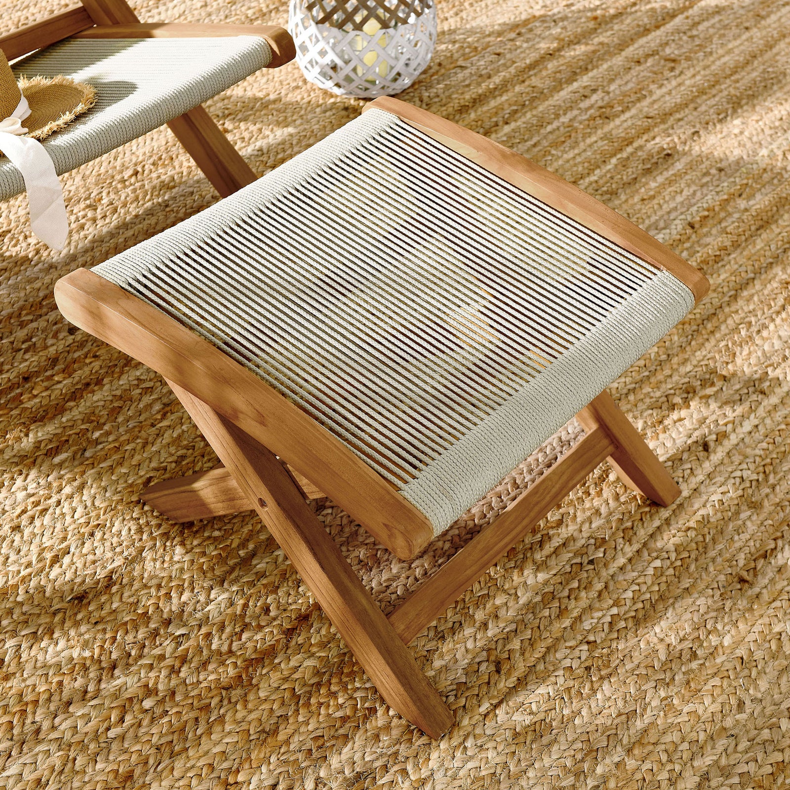 Teak folding ottoman with natural rope seat styled on a woven jute rug for outdoor lounging || EEI-6809-NAT-NAT || front_angle_room