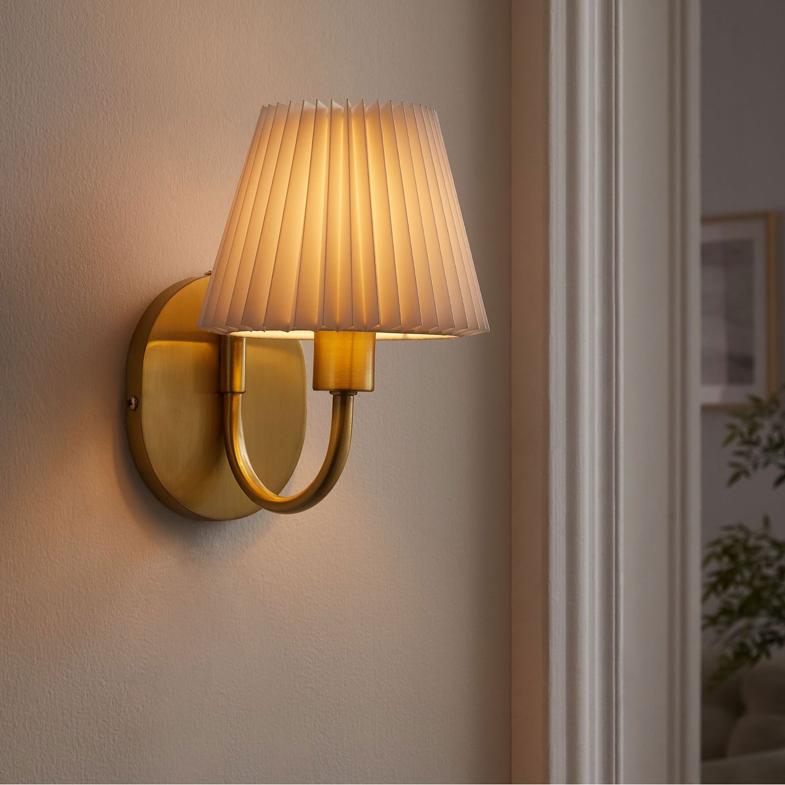 Lit brass U-shaped wall sconce with pleated shade casts a warm glow beside a doorway in the living room. || EEI-6872-BRA || front_angle_on_room