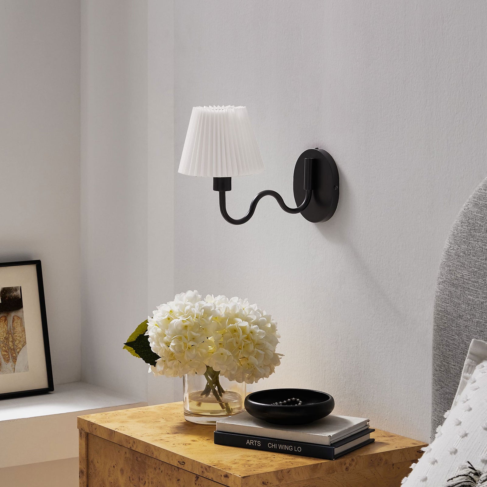 Black squiggle arm wall sconce sits above a bedside table, adding tailored contrast against a bright white wall. || EEI-6873-BLK || front_angle_room