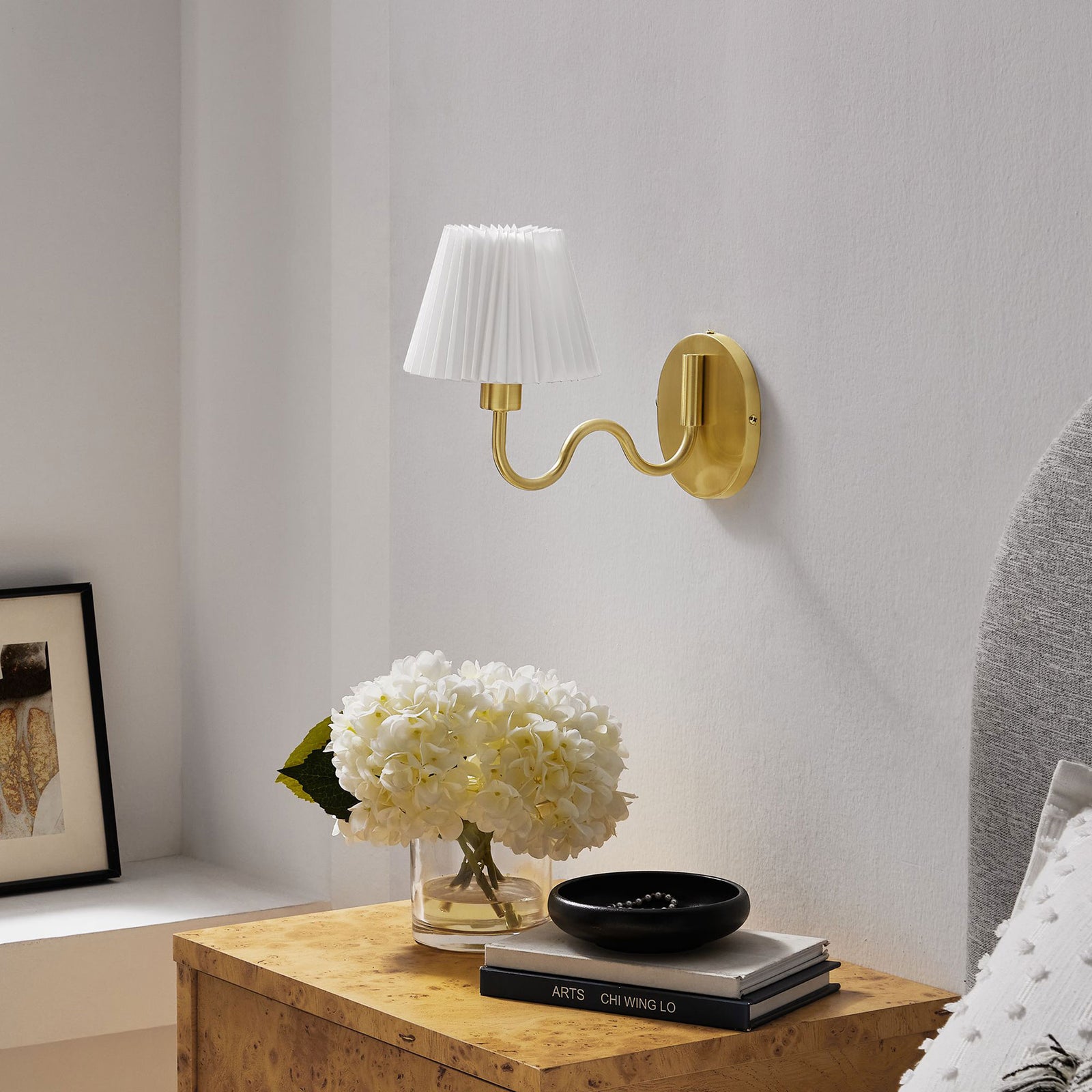 Brass squiggle arm wall sconce stands out against a white wall, adding polished contrast over the bedside table. || EEI-6873-BRA || front_angle_room