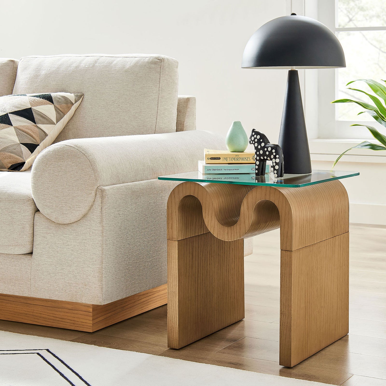Natural Ondine side table styled beside a modern sofa, topped with decor and a lamp on the clear glass surface || EEI-6902-NAT || front_angle_room