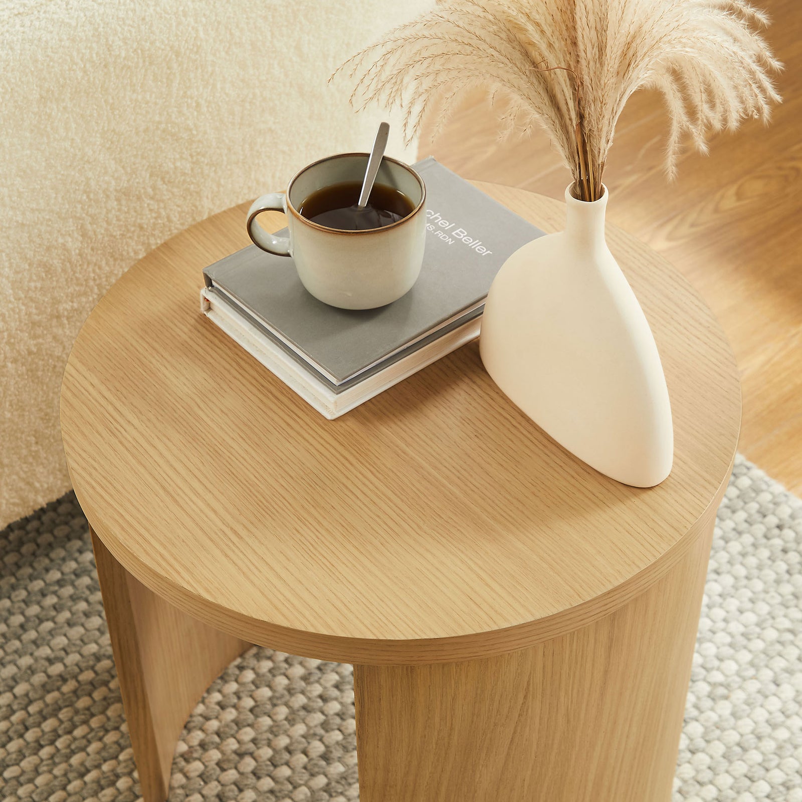 Oak round end table styled with a mug, book, and vase on a textured neutral rug || EEI-6913-OAK || front_angle_room