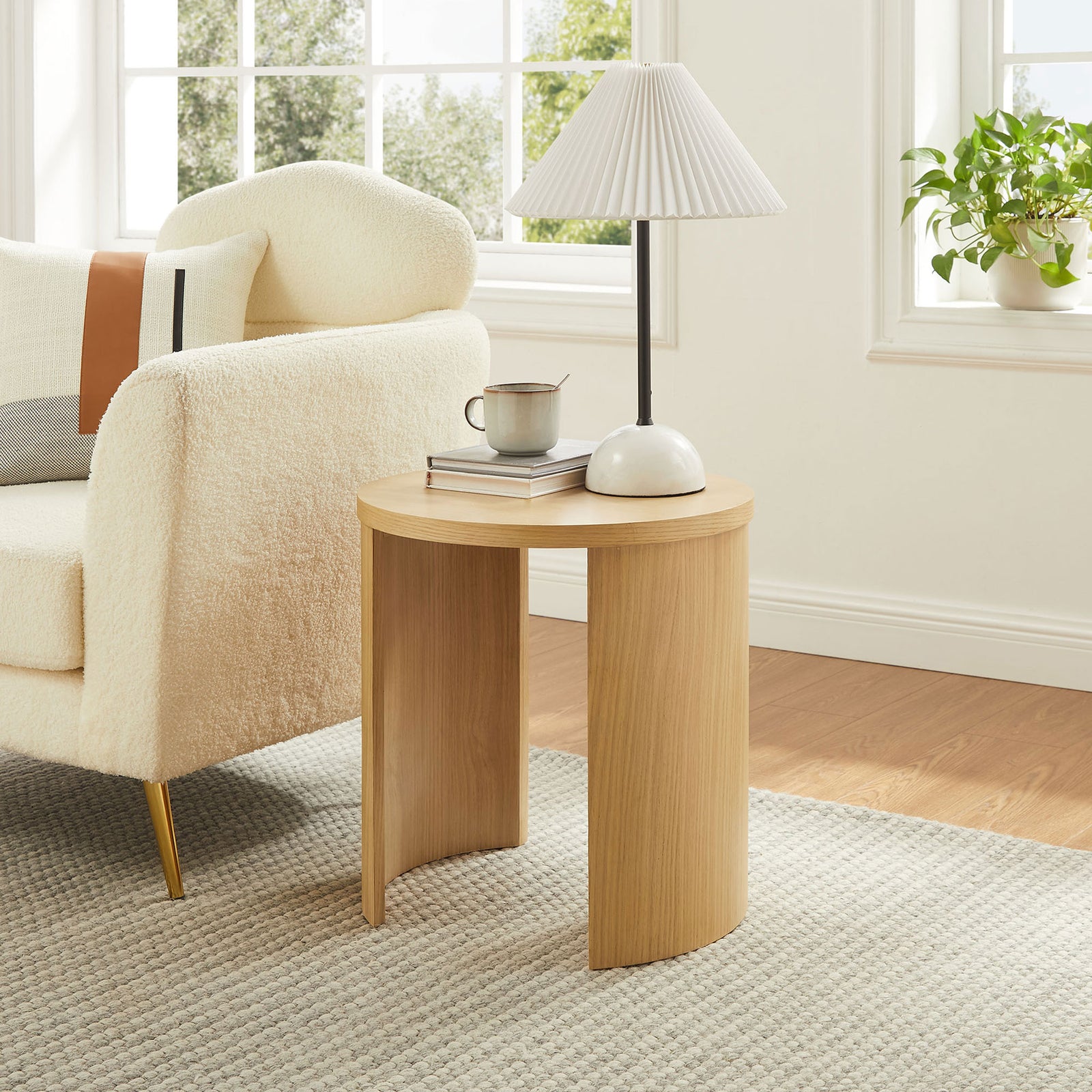Oak round end table beside a cream sofa, topped with a lamp and books in a bright living room || EEI-6913-OAK || front_angle_room
