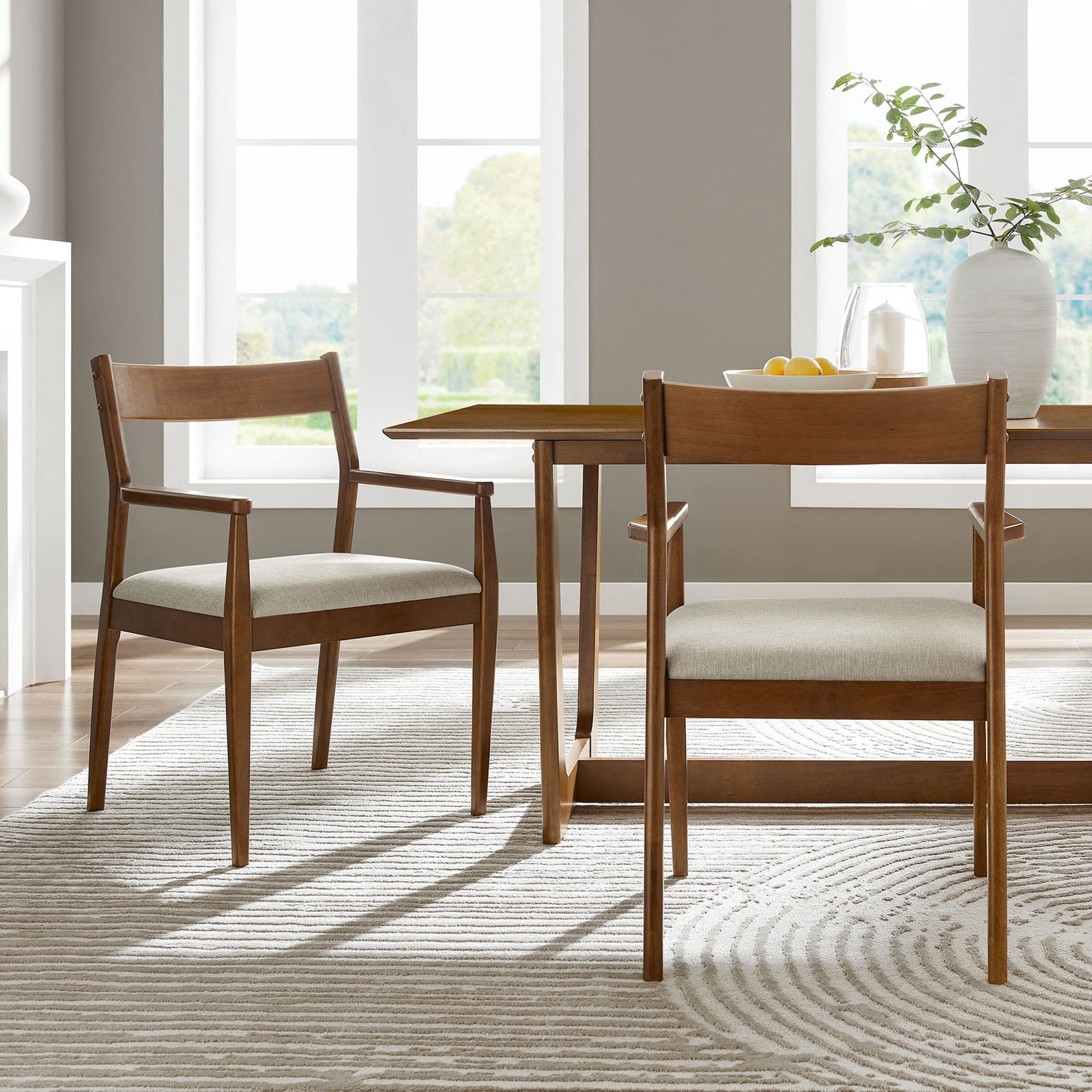 Pair of walnut dining armchairs with beige seats arranged around a rectangular wood table in a bright dining room || EEI-6916-WAL-HEB || front_angle_multi_room