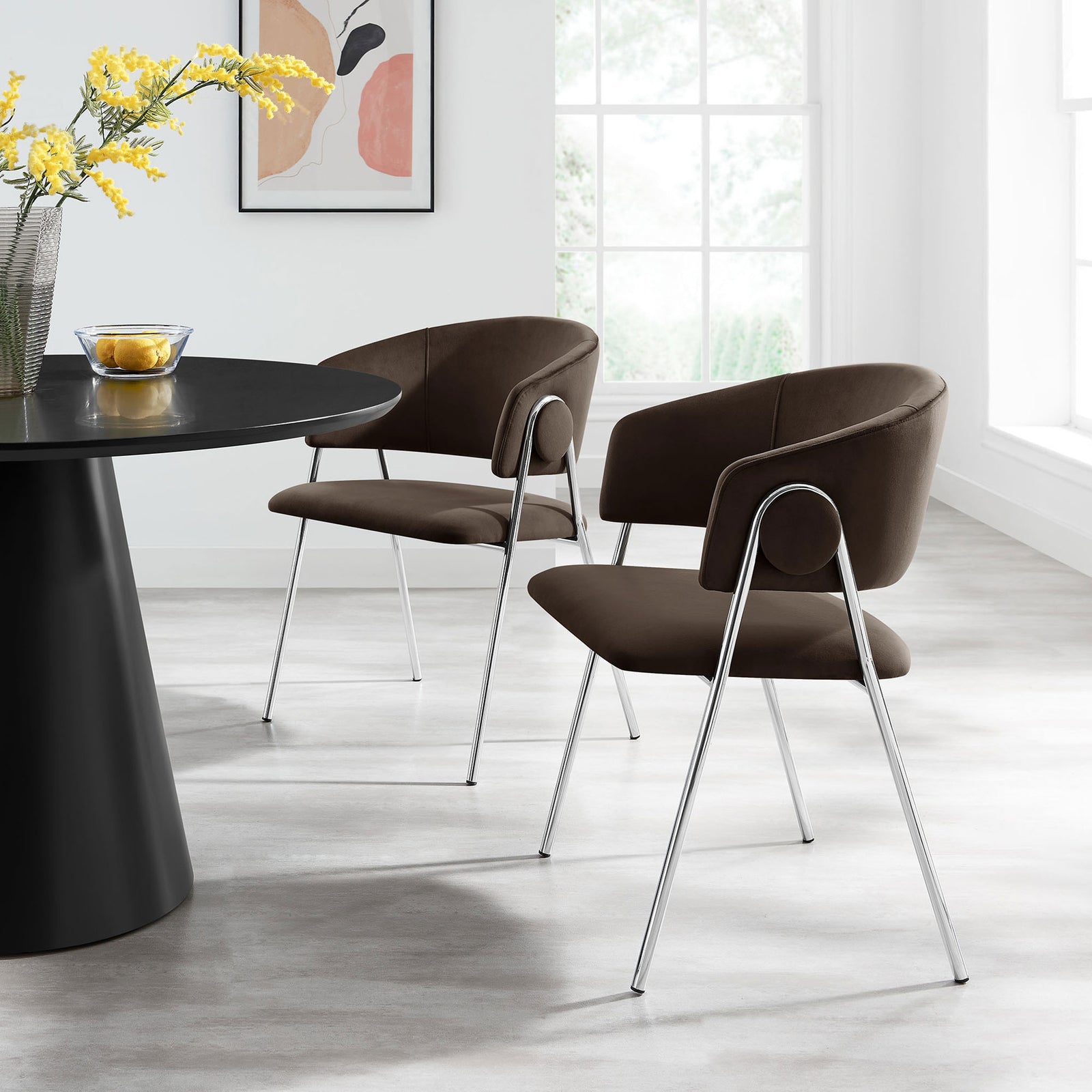 Two Nella velvet dining chairs with chrome legs arranged around a modern round table in a bright dining room. || EEI-7046-CHB-SLV || front_angle_multi_room