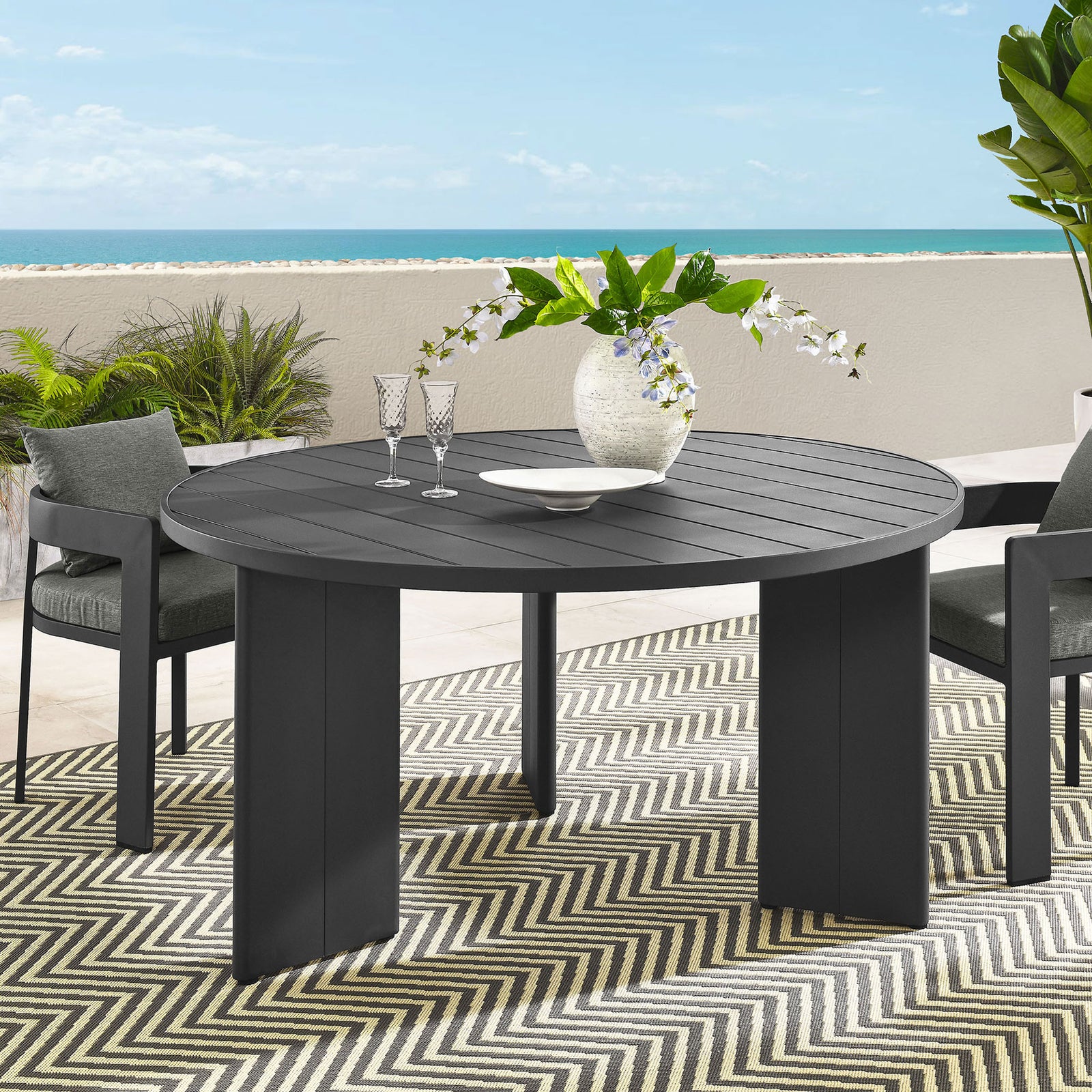 Tahoe round patio table styled with lounge chairs, rug, and greenery for relaxed outdoor dining. || EEI-7055-GRY || front_angle_room