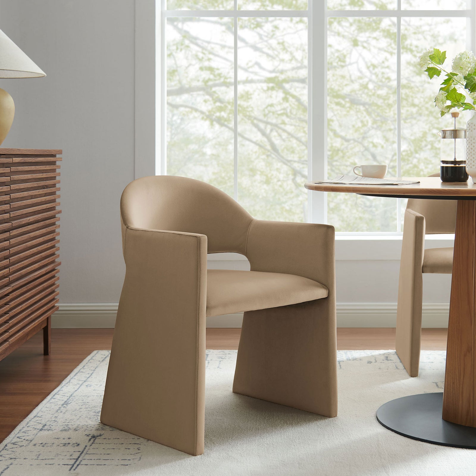 Taupe velvet dining armchair styled in a bright dining room beside a round wood table and sideboard || EEI-7081-TAU || front_angle_room