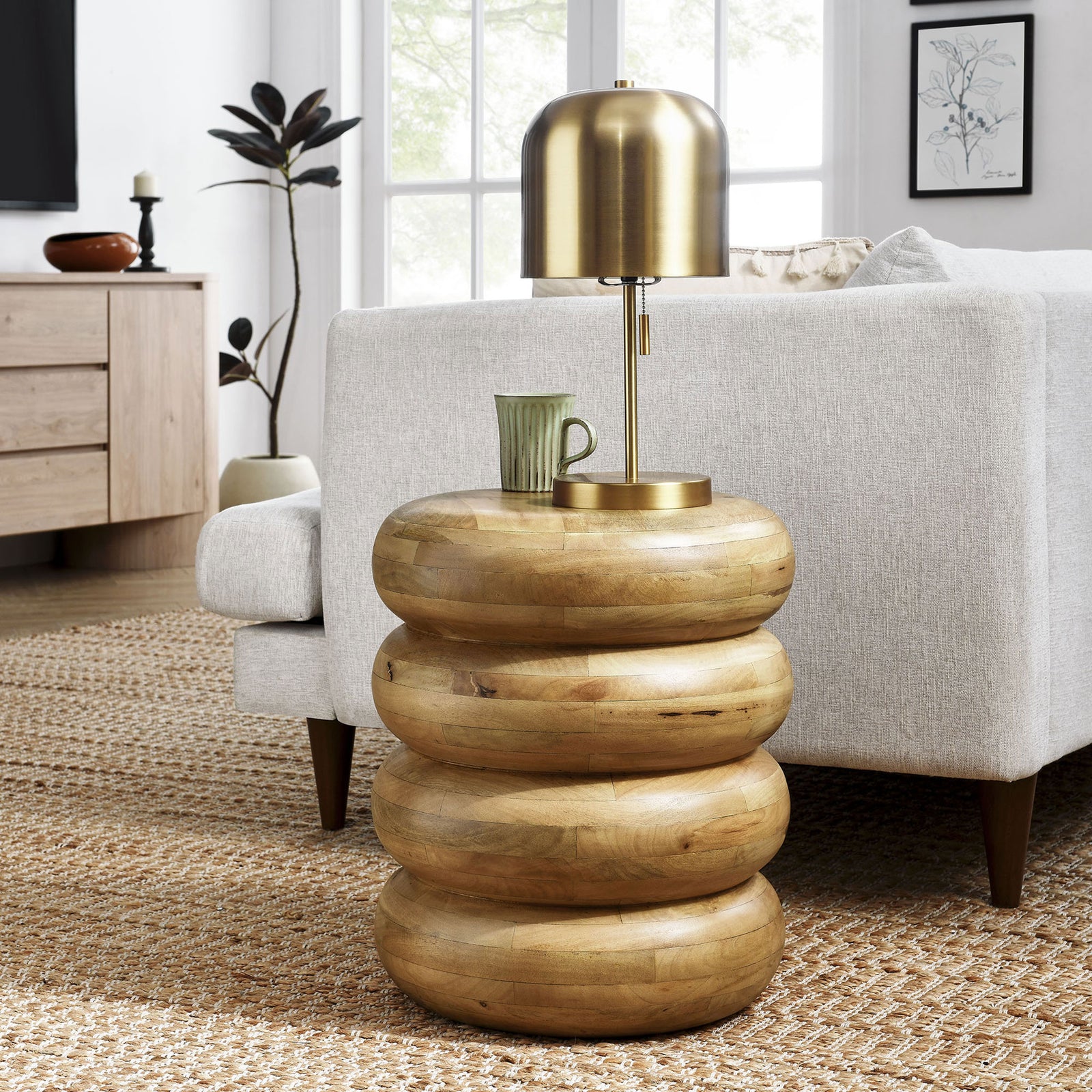 Natural stacked wood side table beside a modern sofa, topped with a brass lamp and mug || EEI-7106-NAT || front_room