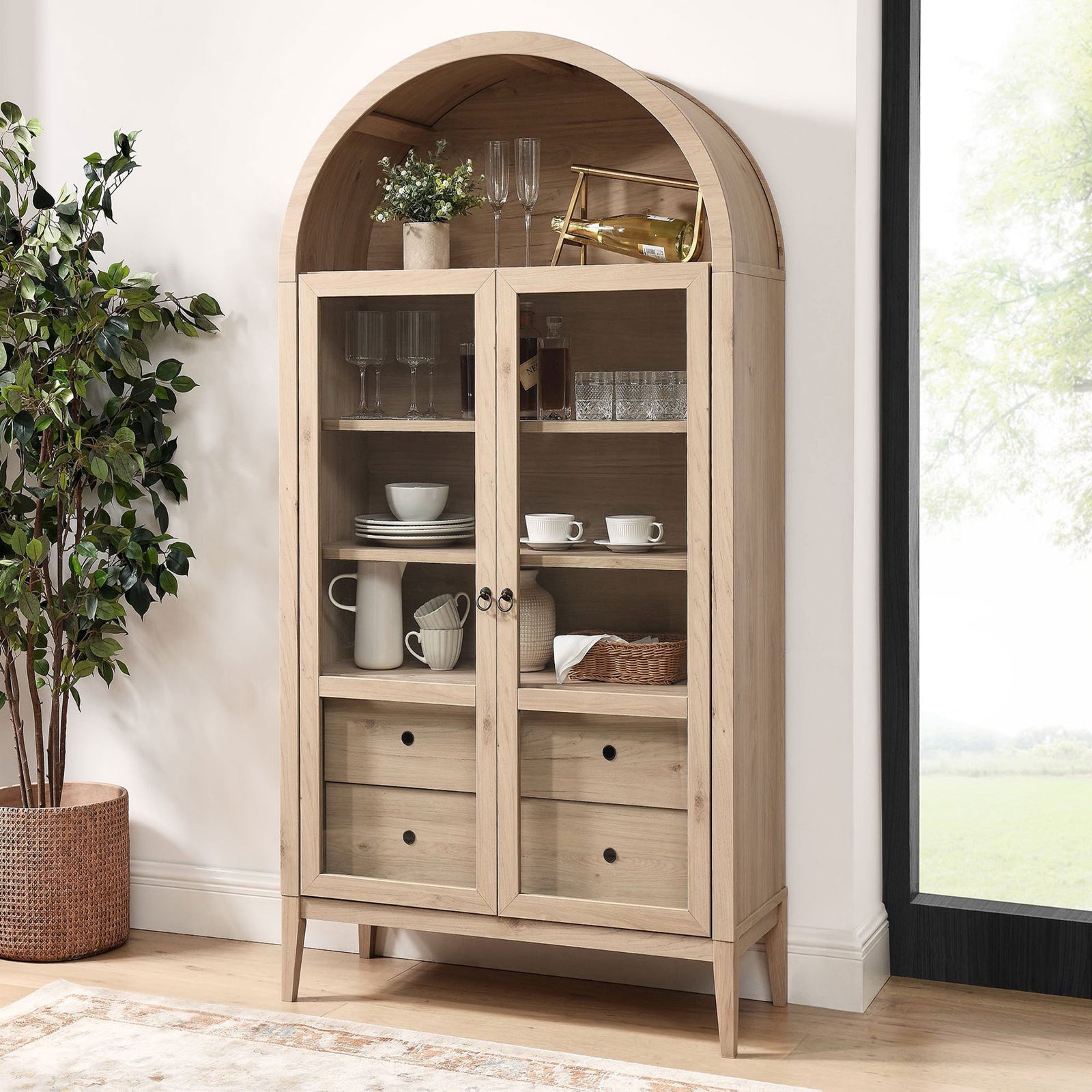 Light oak display cabinet filled with glassware and bar accessories beside a sunlit window || EEI-7168-LOA || front_angle_room