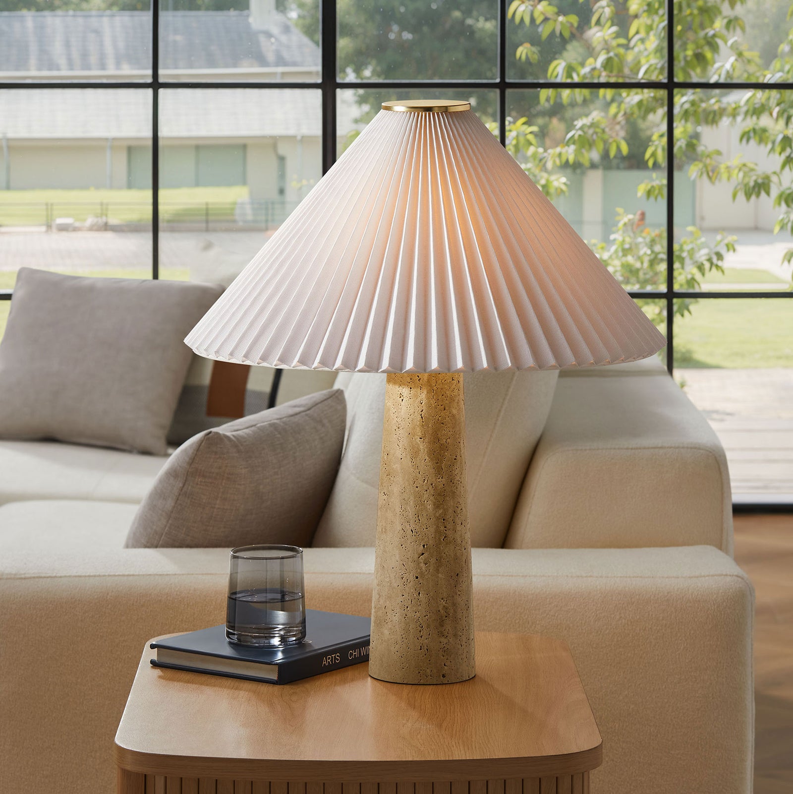 Lamp casts a warm glow through the pleated shade, highlighting the travertine base beside a modern cream sofa || EEI-7215-TRV || front_on_room
