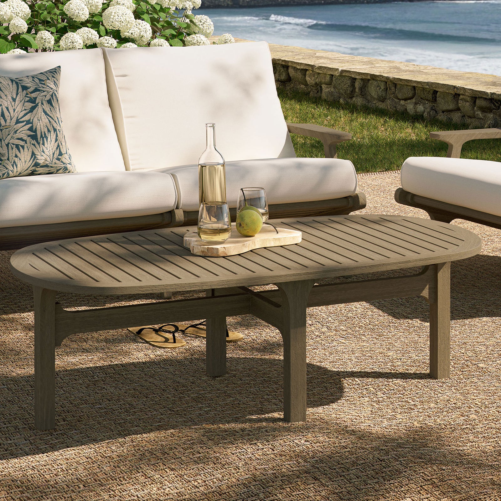 Saratoga oval outdoor teak coffee table in weathered gray styled on a coastal patio seating area || EEI-7246-WGR || front_angle_room