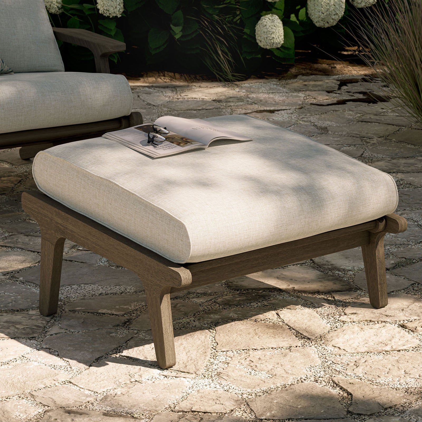 Pebble cushioned teak ottoman styled outdoors as a chic footrest and flexible extra patio seat || EEI-7250-WGR-PEB || front_angle_room