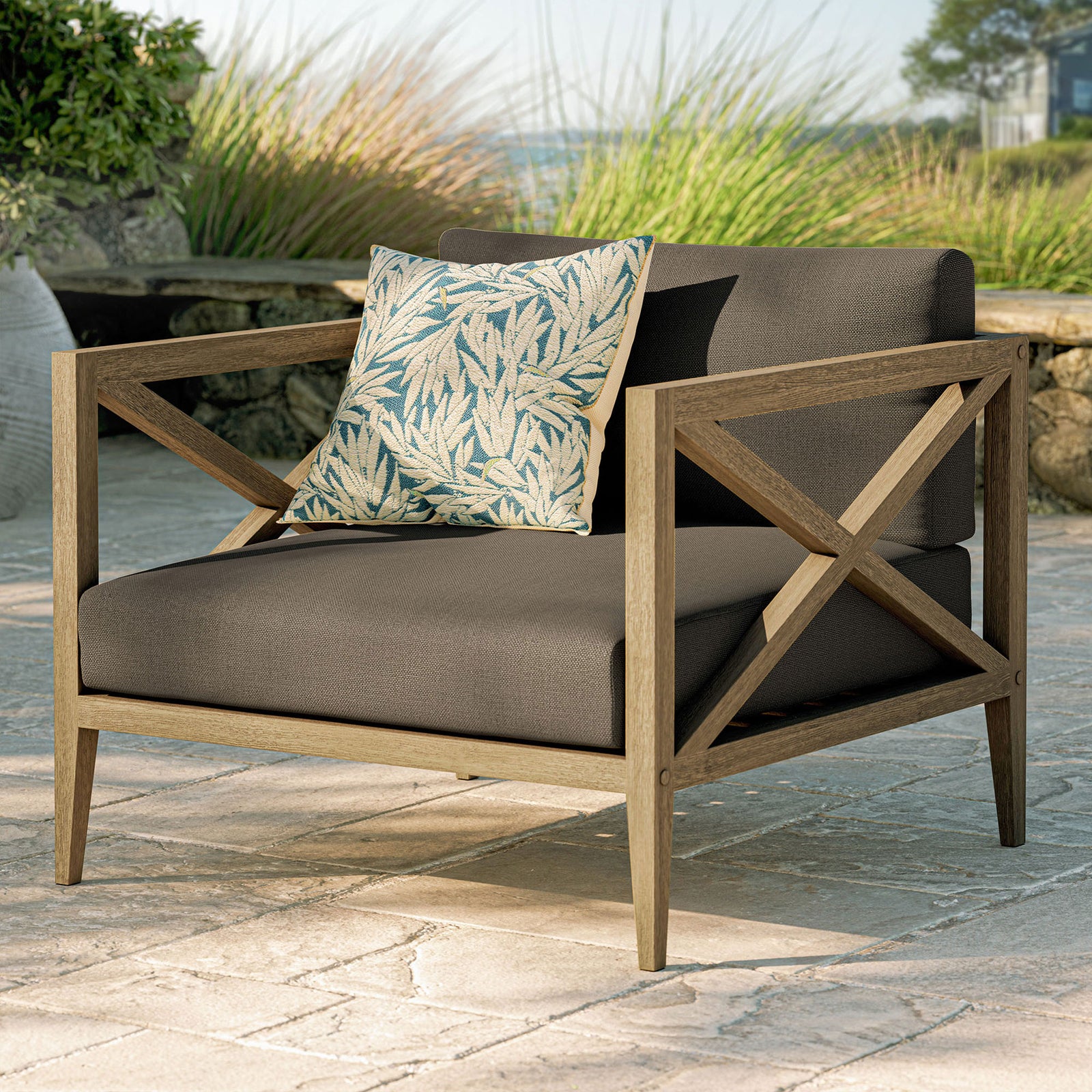 Northlake teak armchair with flint gray cushions styled with a leaf print pillow on an outdoor patio || EEI-7252-WGR-FLI || front_angle_room
