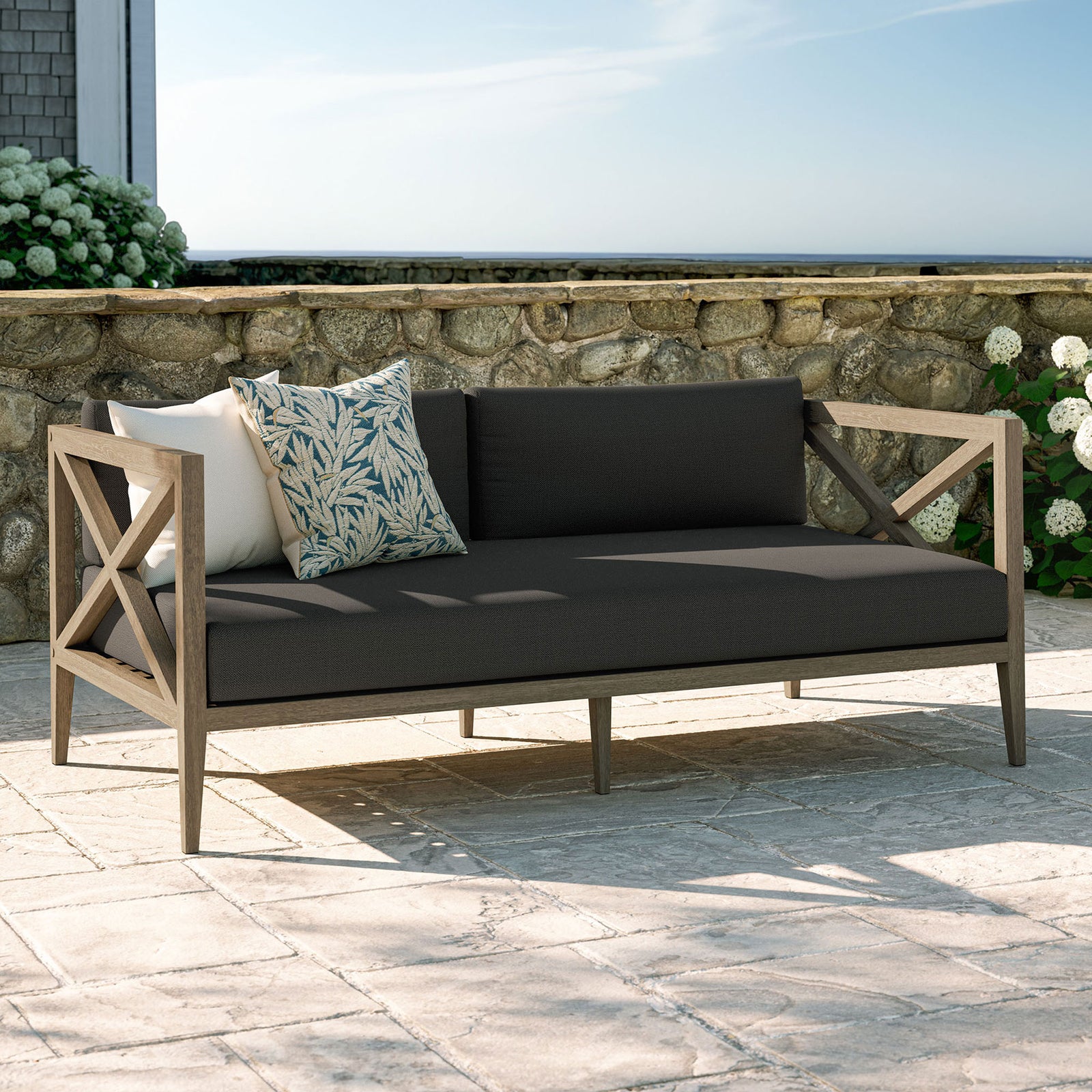 Flint gray cushioned teak loveseat styled on a stone patio, perfect for garden lounges and coastal decks. || EEI-7254-WGR-FLI || front_angle_room