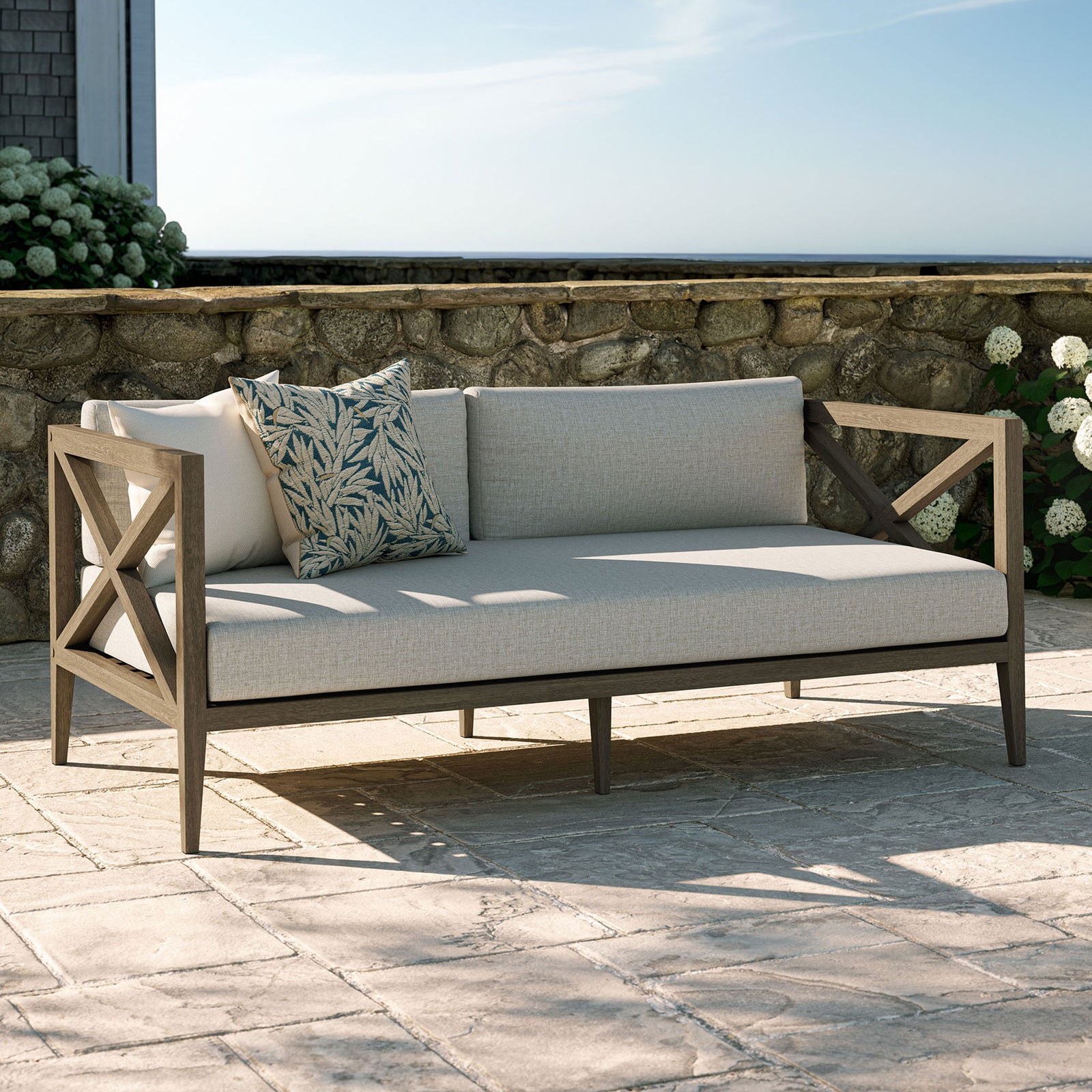 Pebble cushioned teak loveseat styled on a stone patio, perfect for garden lounges and coastal decks. || EEI-7254-WGR-PEB || front_angle_room