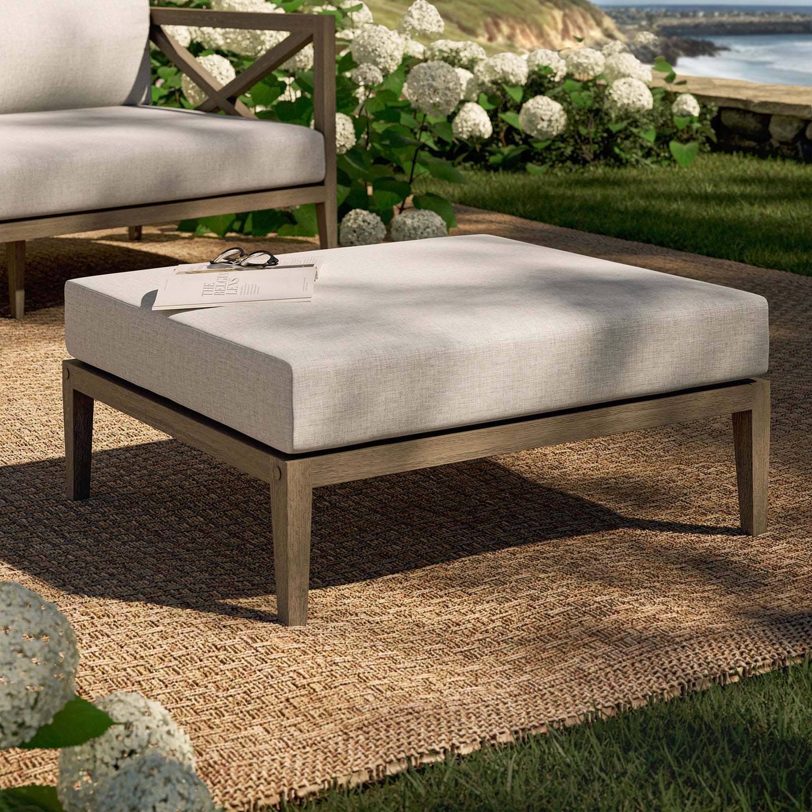 Teak patio ottoman with beige cushion styled on a woven rug amid lush garden greenery || EEI-7255-WGR-PEB || front_angle_room
