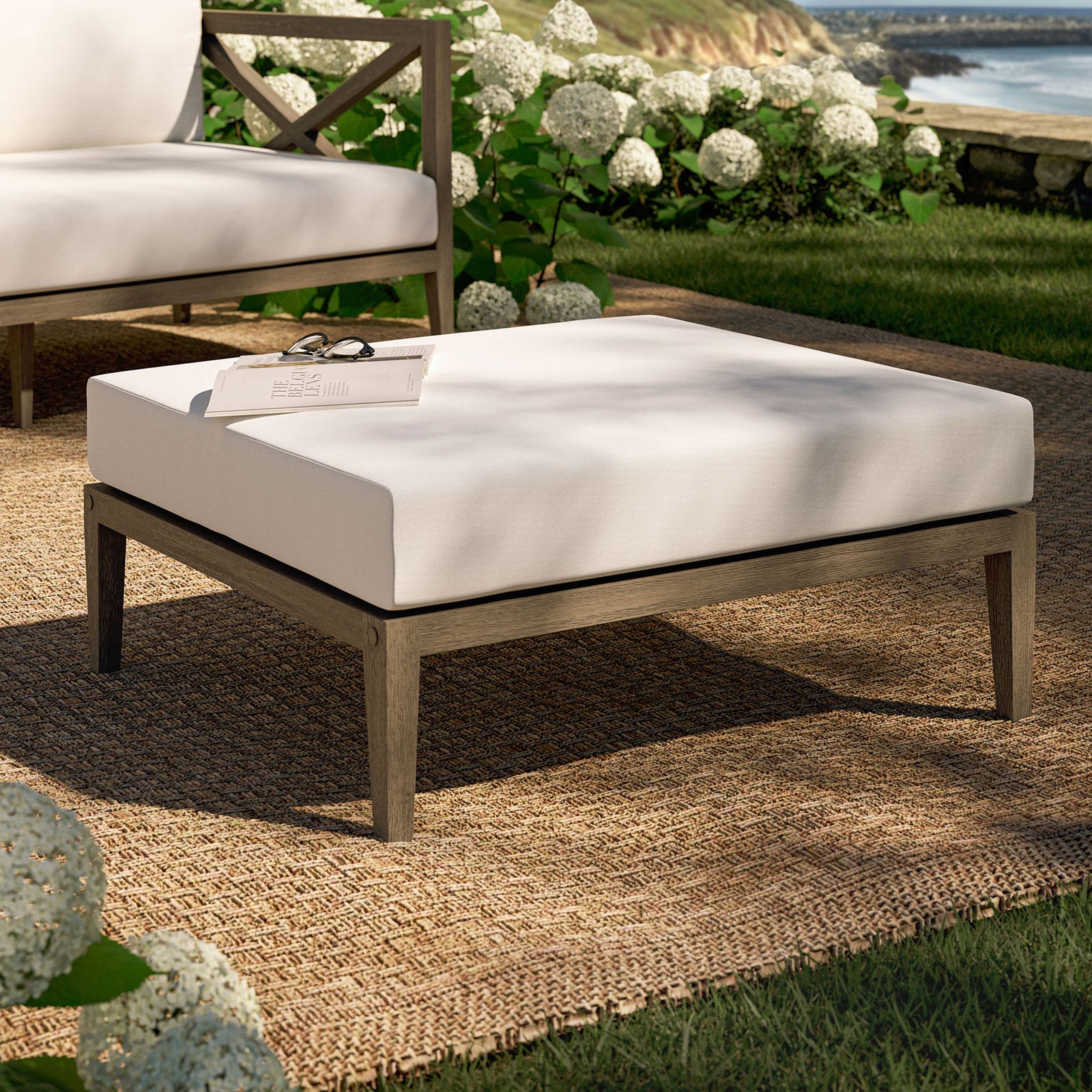 Teak patio ottoman with white cushion styled on a woven rug amid lush garden greenery || EEI-7255-WGR-WHI || front_angle_room